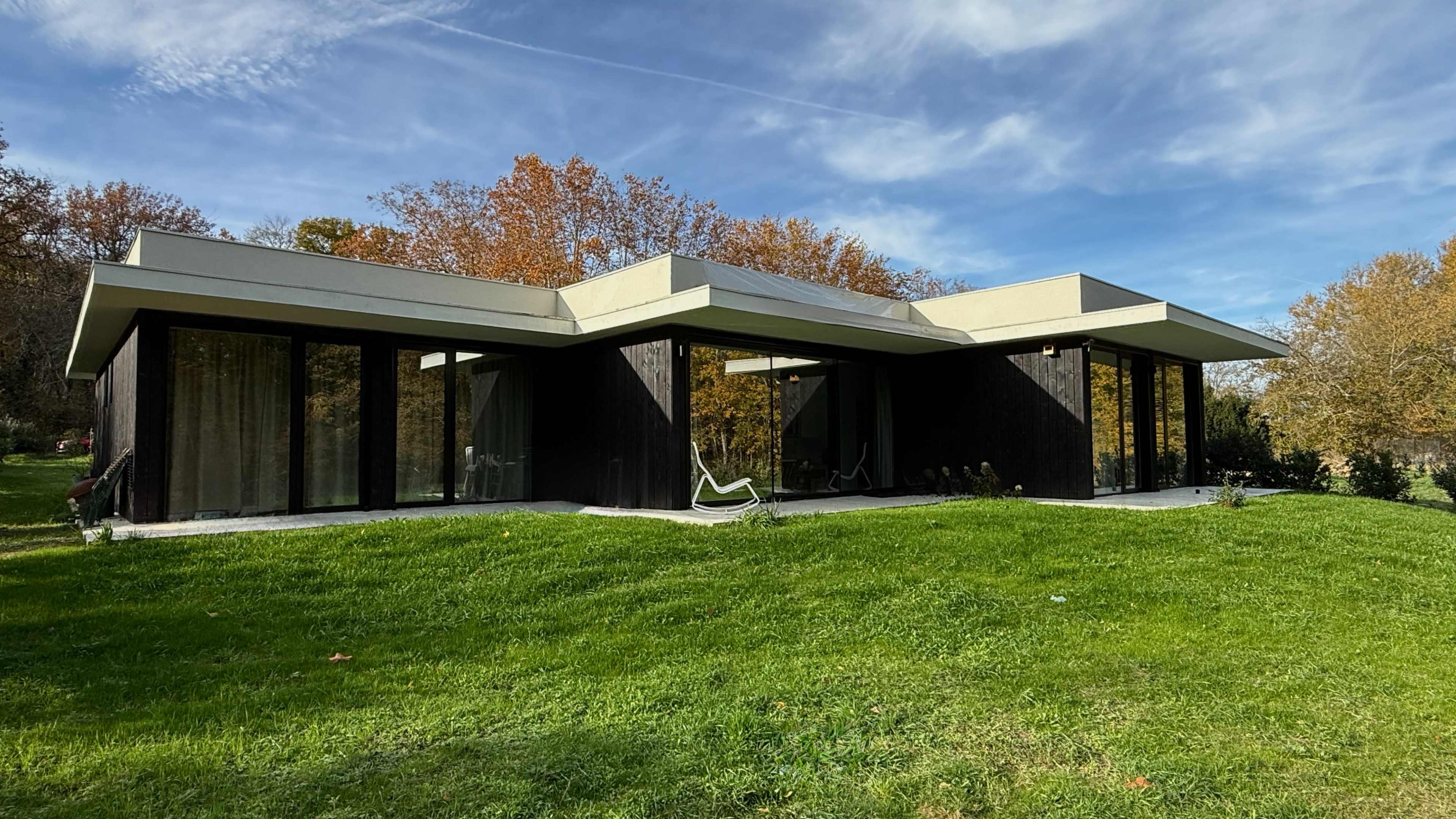 The image shows a modern, single-story house with a flat roof, large glass windows, and a dark exterior set in a grassy area surrounded by trees with autumn foliage.