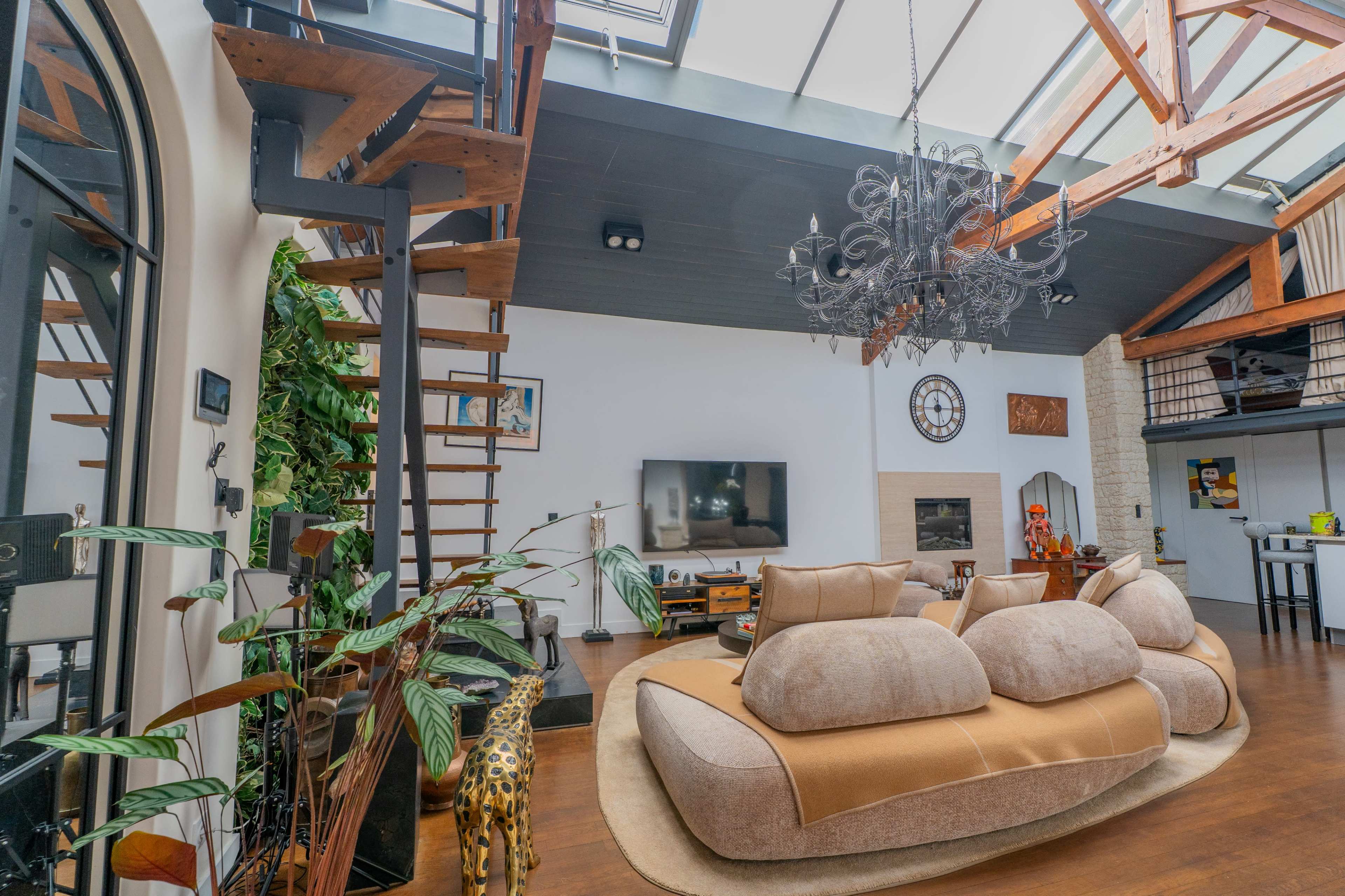 The image shows a spacious living room featuring large windows, a modern chandelier, a spiral staircase, a central sofa, and decorative plants.