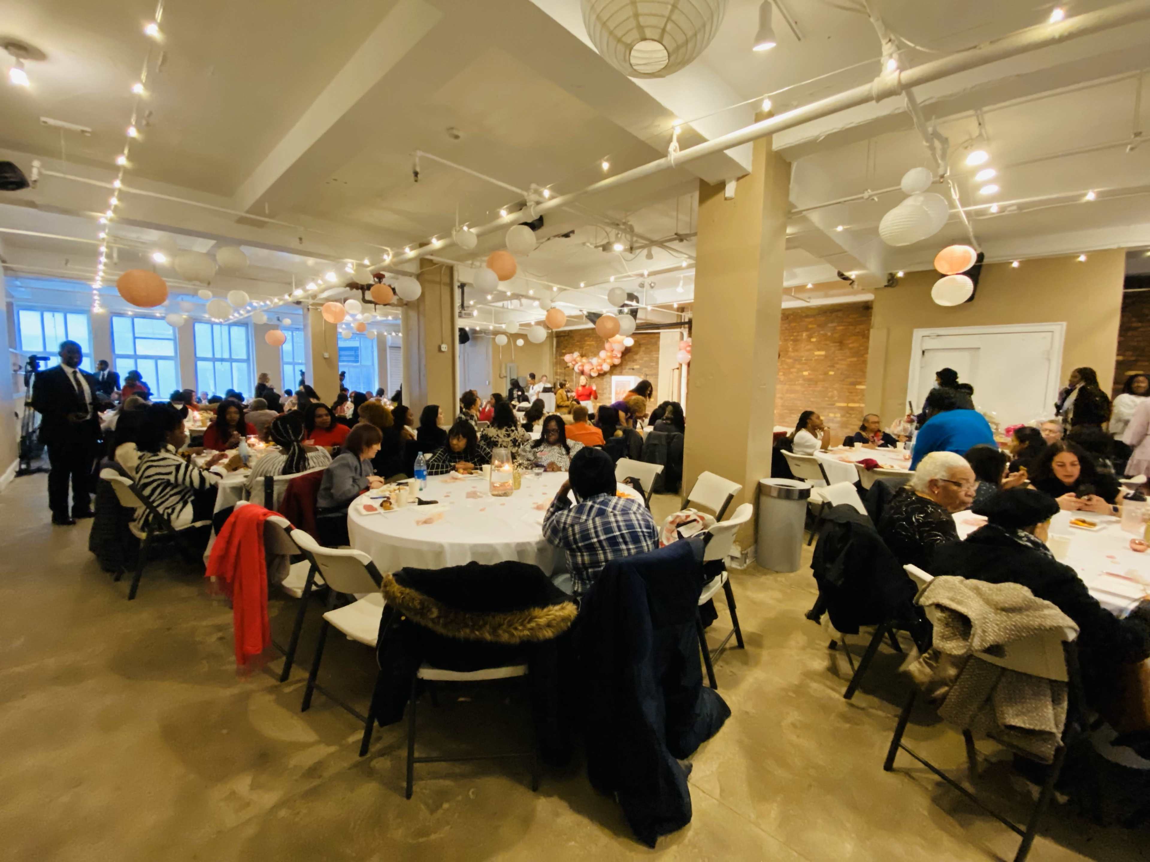 A large indoor event space is filled with round tables where many people are seated, dining and socializing under decorative lighting and hanging lanterns.