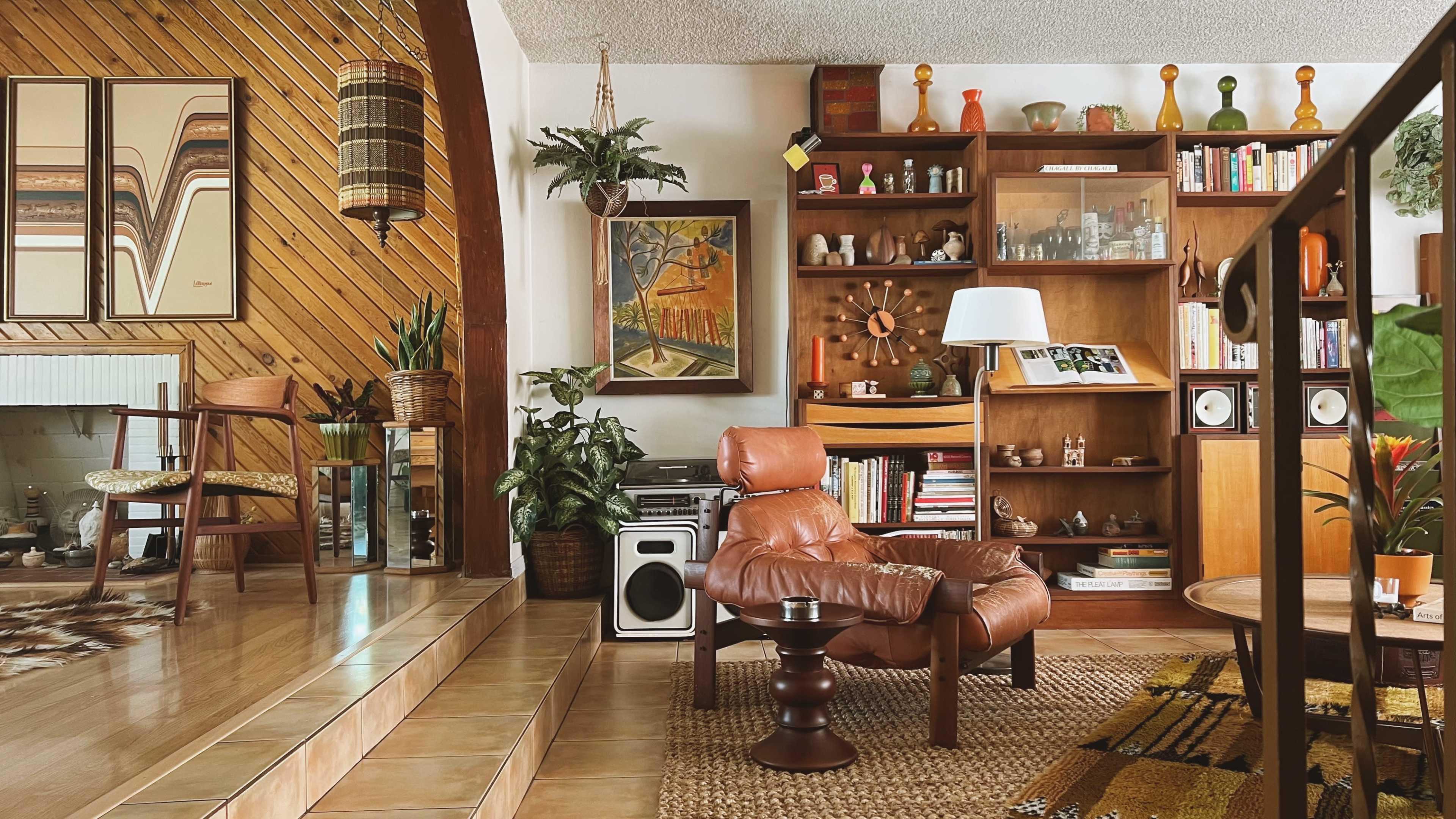 A cozy living room with wooden paneling, a brown leather chair, a bookshelf filled with various books and decor items, and green plants scattered throughout the space.