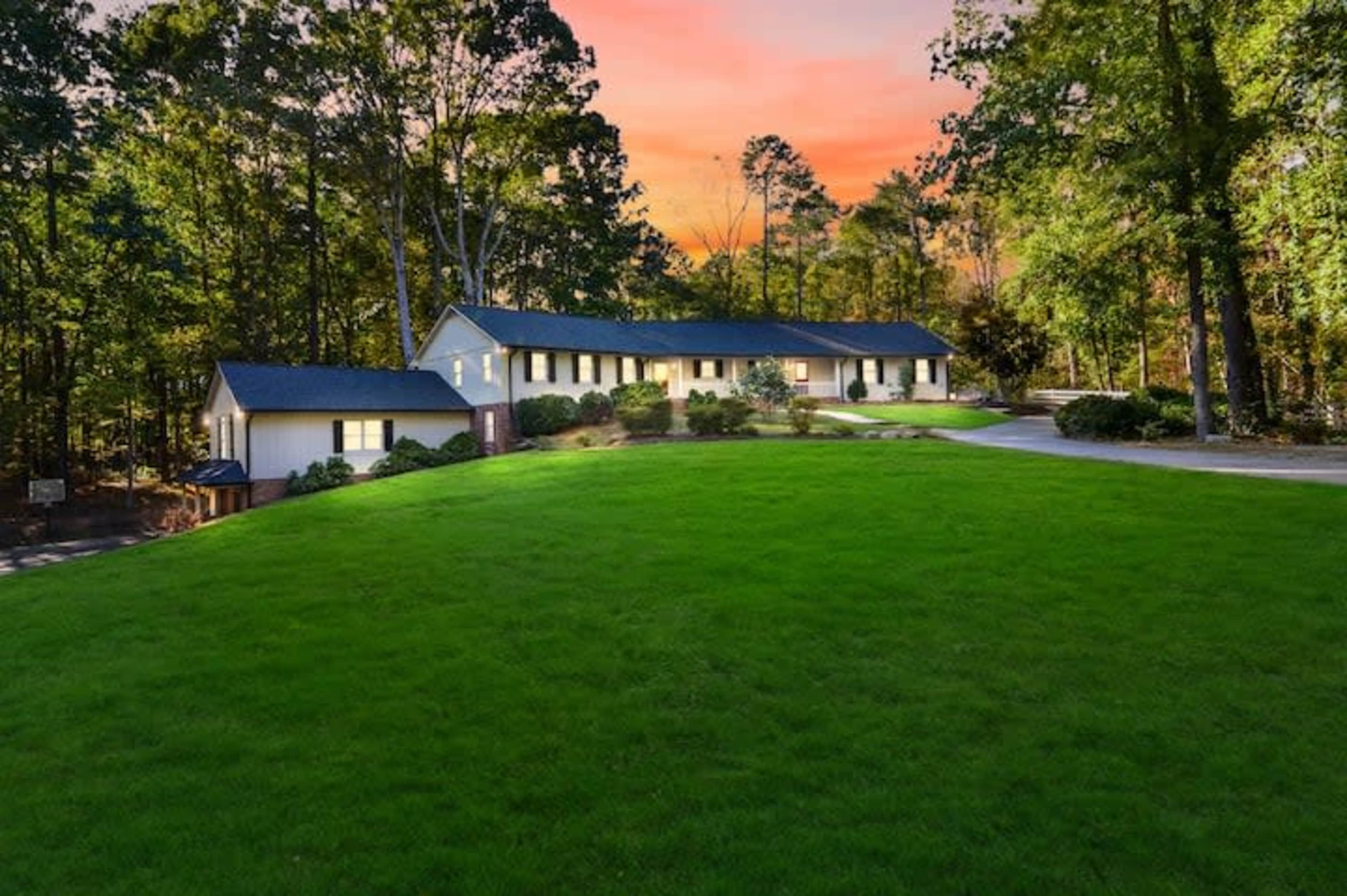 A single-story house is situated on a lush, green lawn surrounded by trees, with a sunset illuminating the sky in the background.