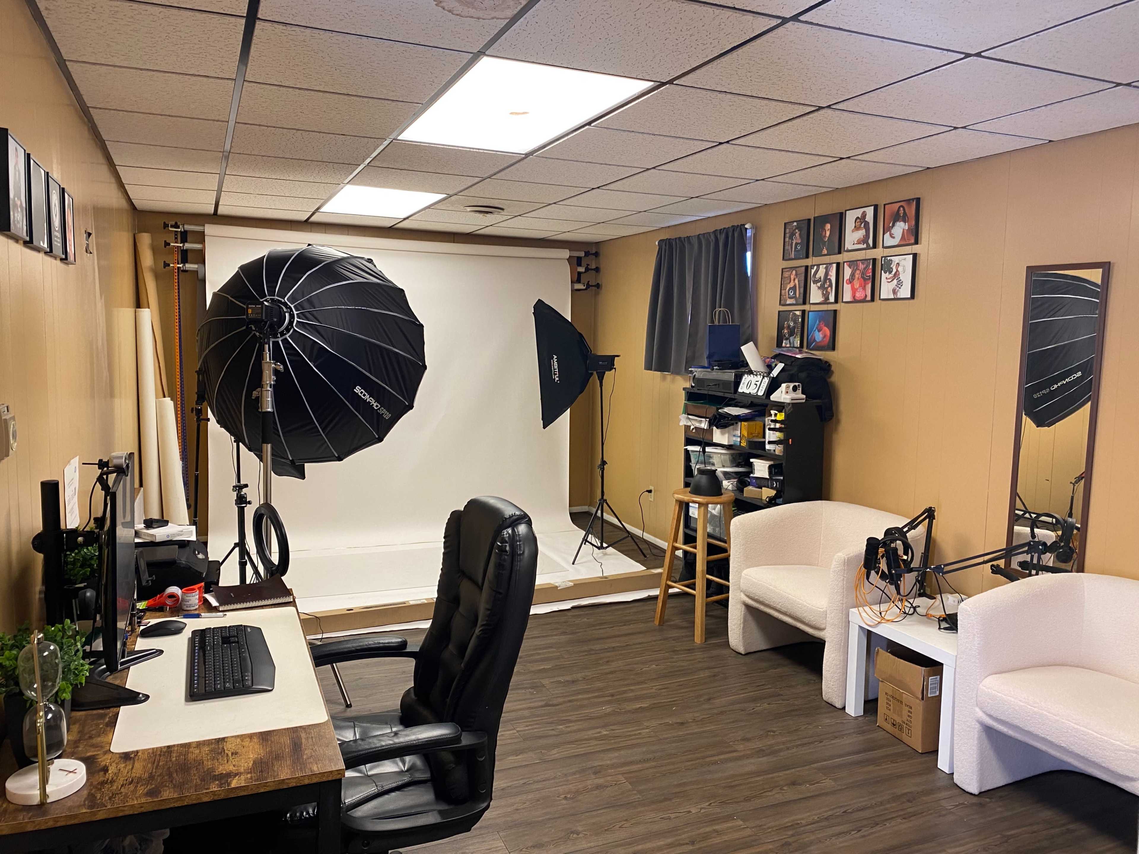 The image shows a photography studio with a computer desk, two large softbox lights, a backdrop, and seating areas.