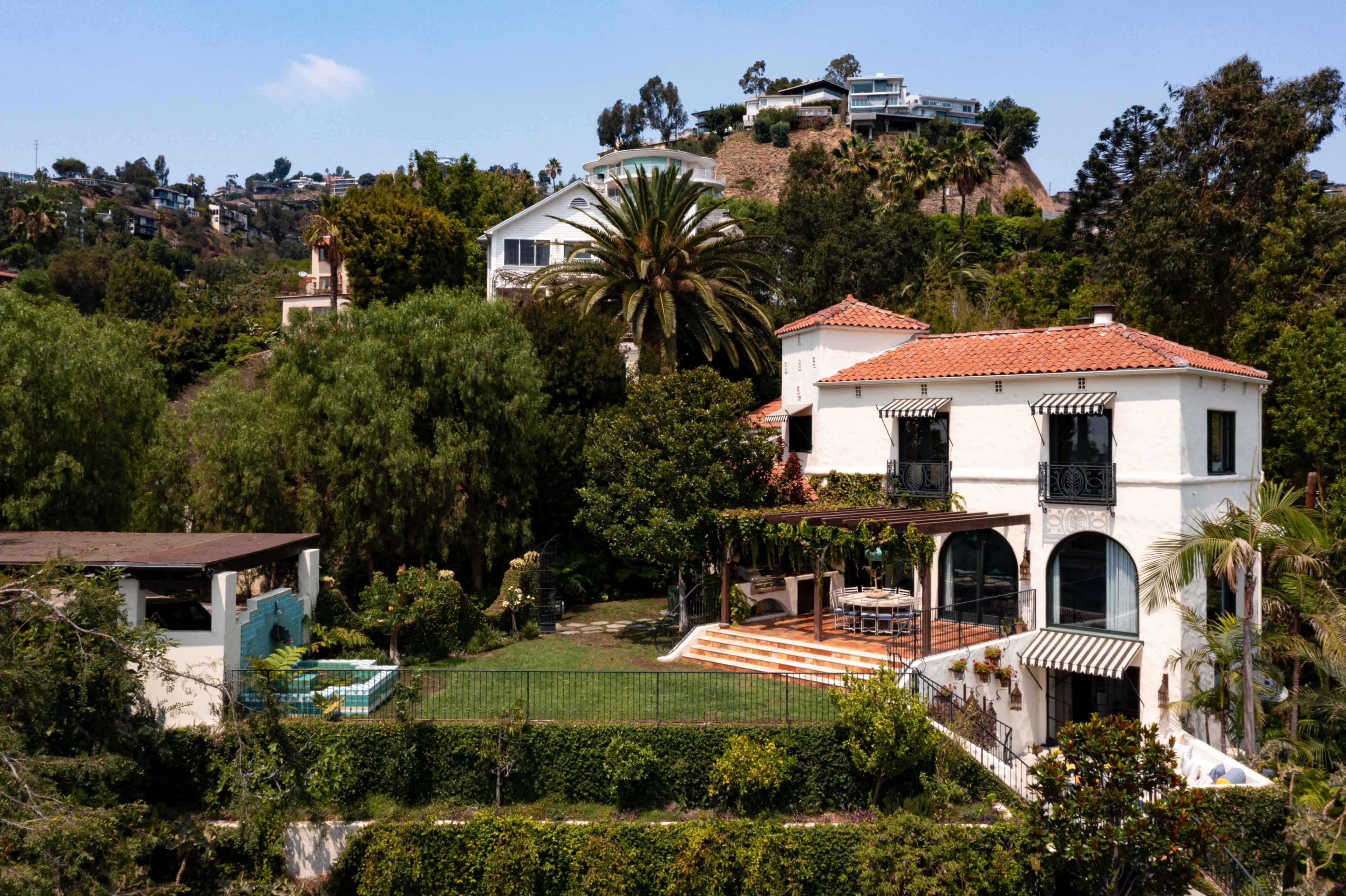The image shows a white house with a red-tiled roof and a large patio, surrounded by greenery and other buildings on a hillside.