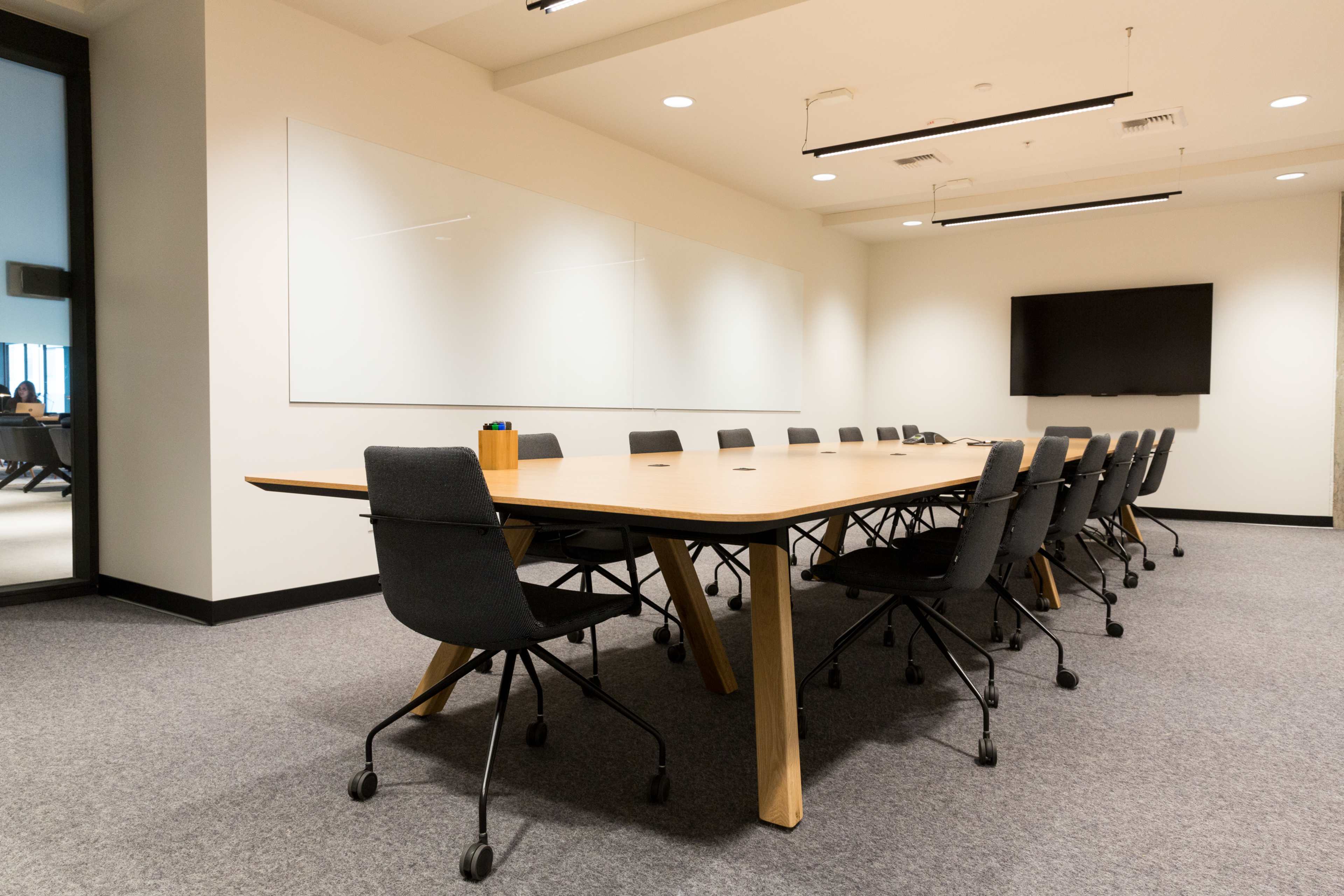 A large conference room features a long wooden table surrounded by black rolling chairs and a wall-mounted television.