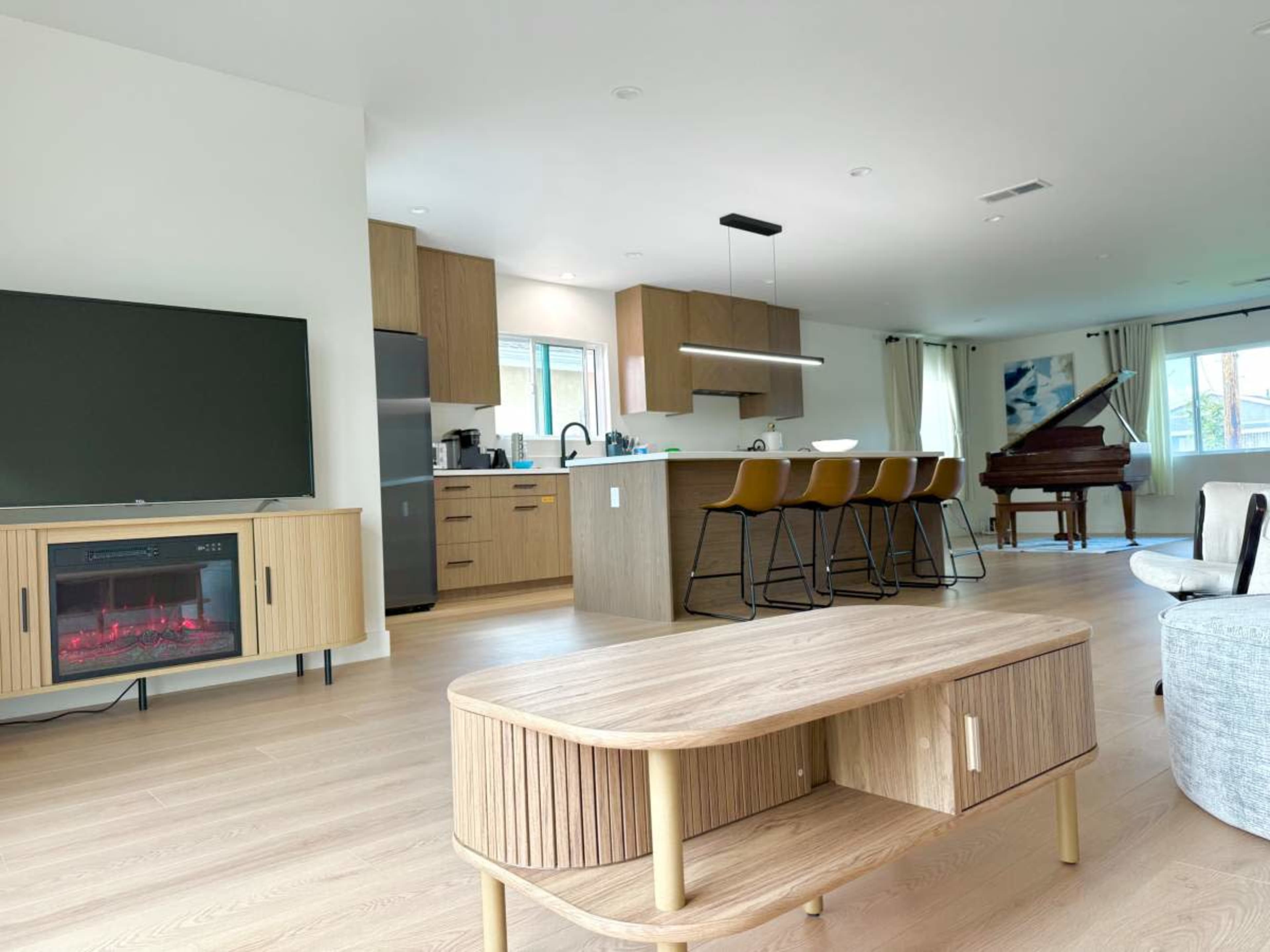 The image shows a modern, open-concept living space featuring a kitchen with bar seating, a dining area, and a grand piano.