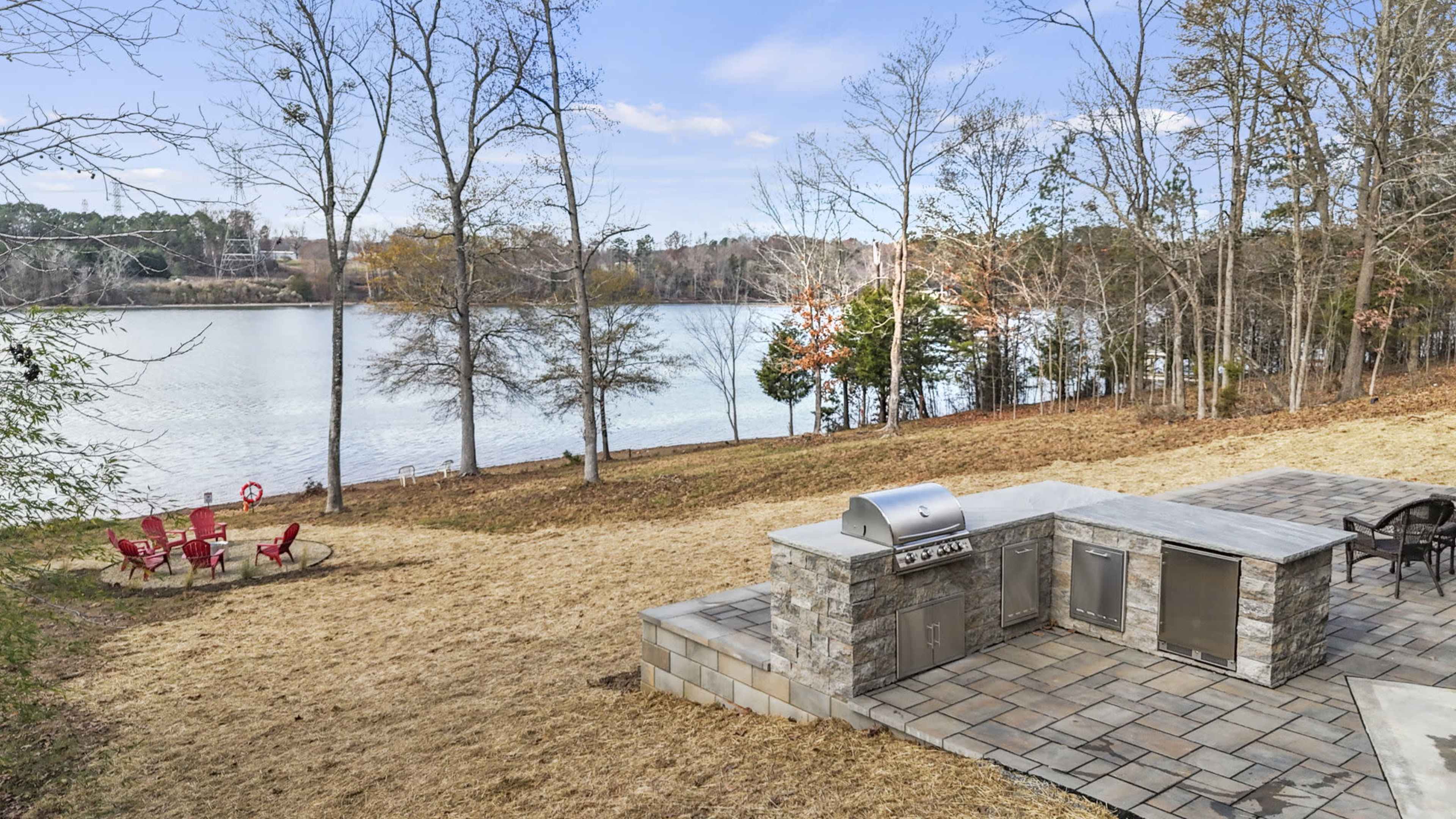 The image shows a lakeside patio with a stone outdoor kitchen area and several red Adirondack chairs arranged on a grassy bank.