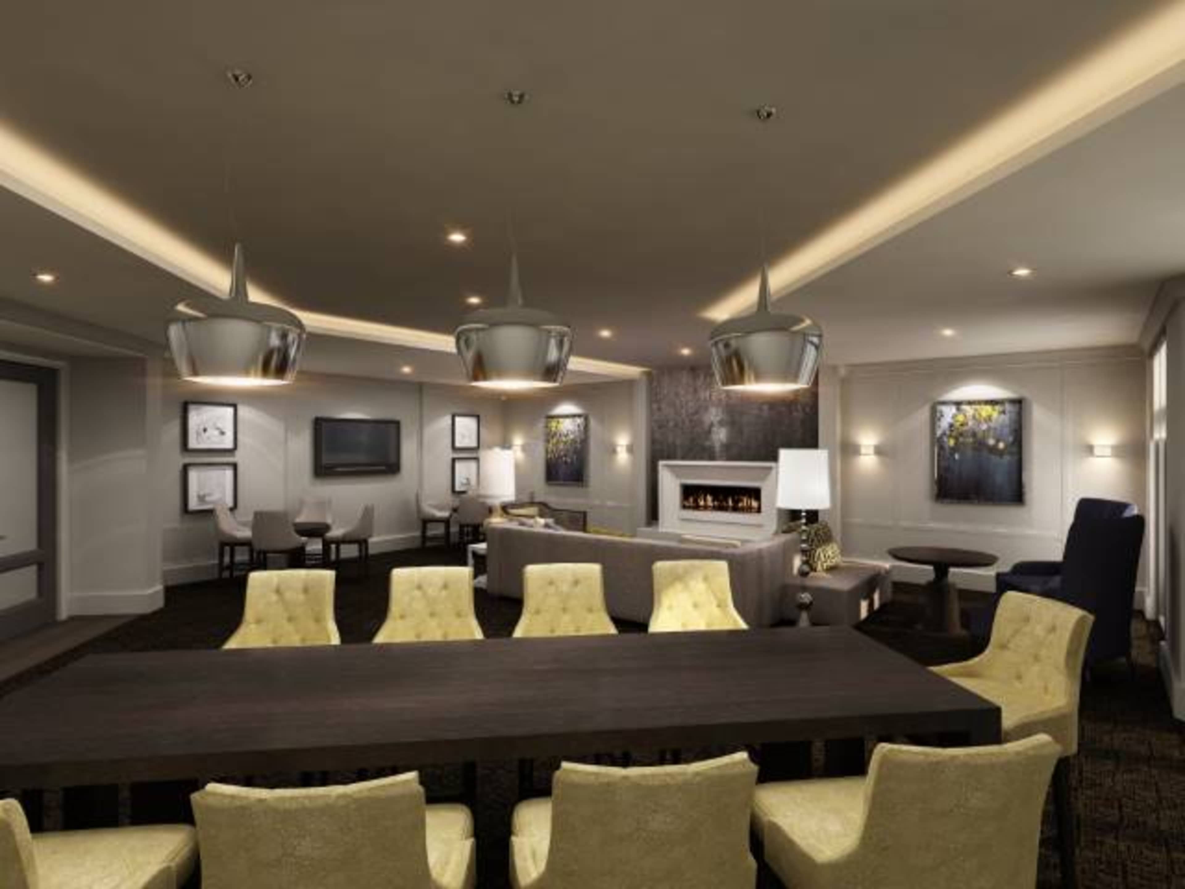 A modern lounge area with a long wooden dining table surrounded by yellow chairs, a fireplace, and various seating arrangements under sleek pendant lights.