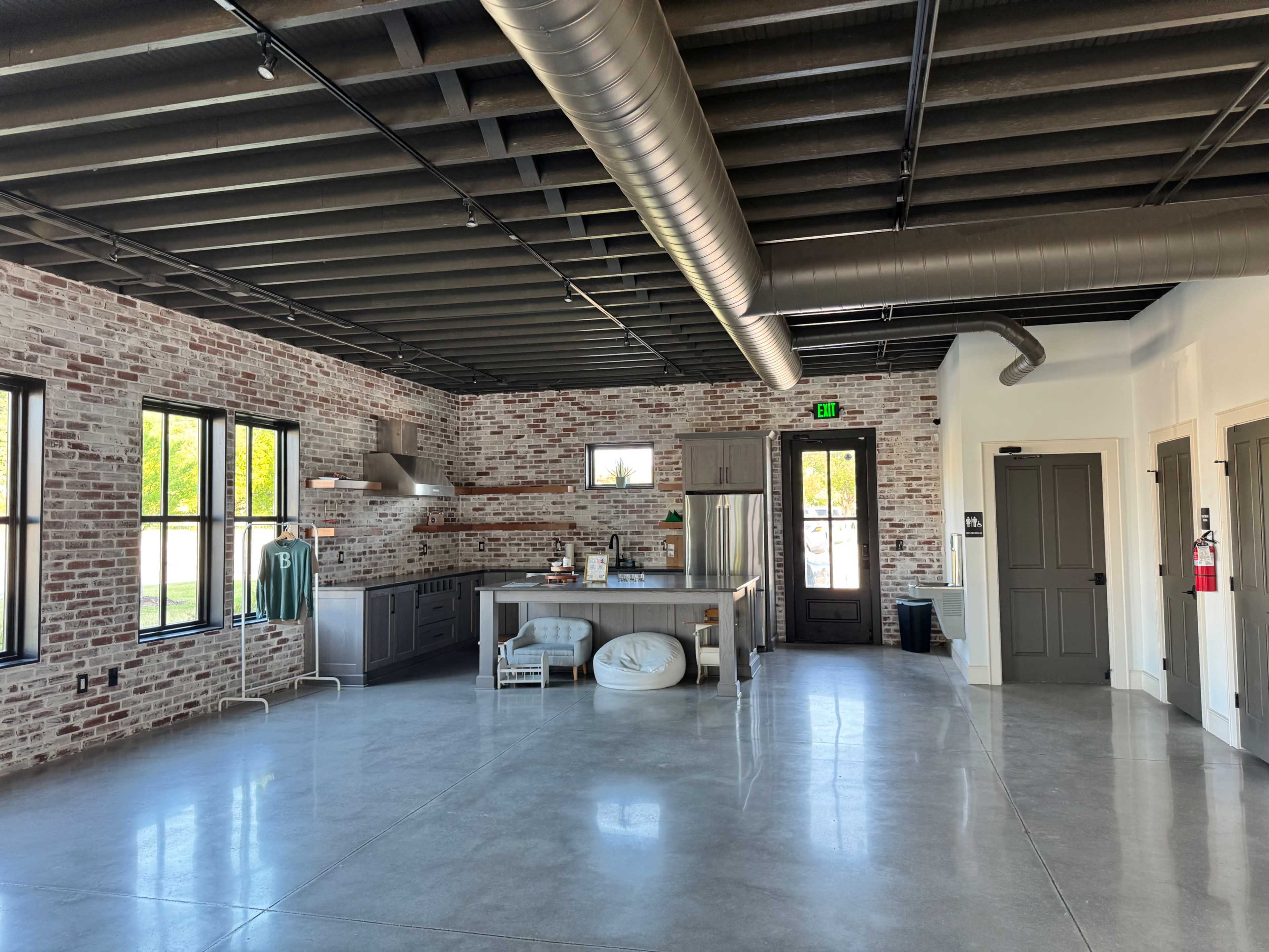 The image shows an open interior space with exposed brick walls, a modern kitchen area, large windows, and polished concrete flooring.