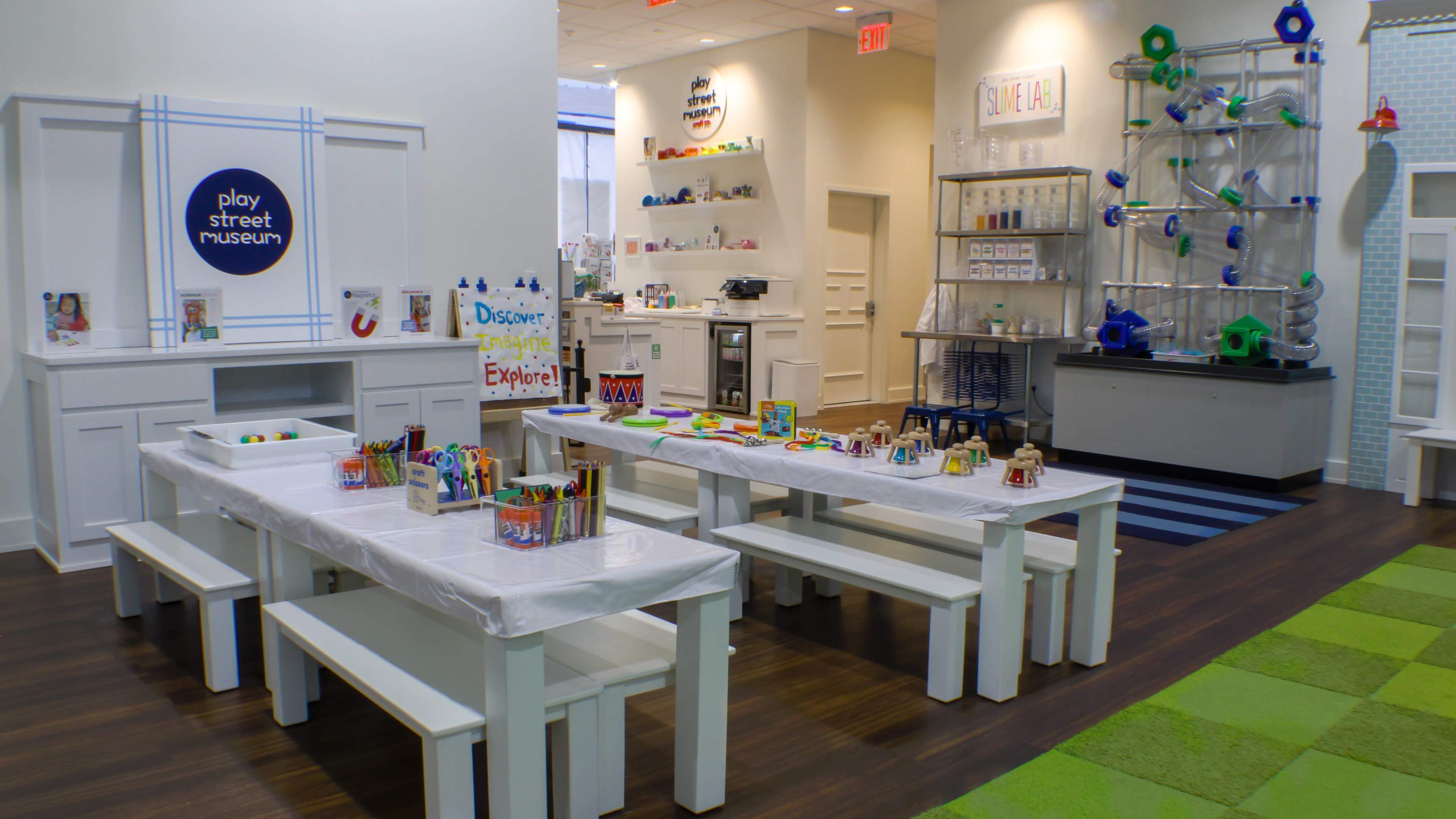 A brightly lit children's museum space featuring several white tables equipped with art supplies, a play area with colorful interactive structures, and a clean, organized backdrop.