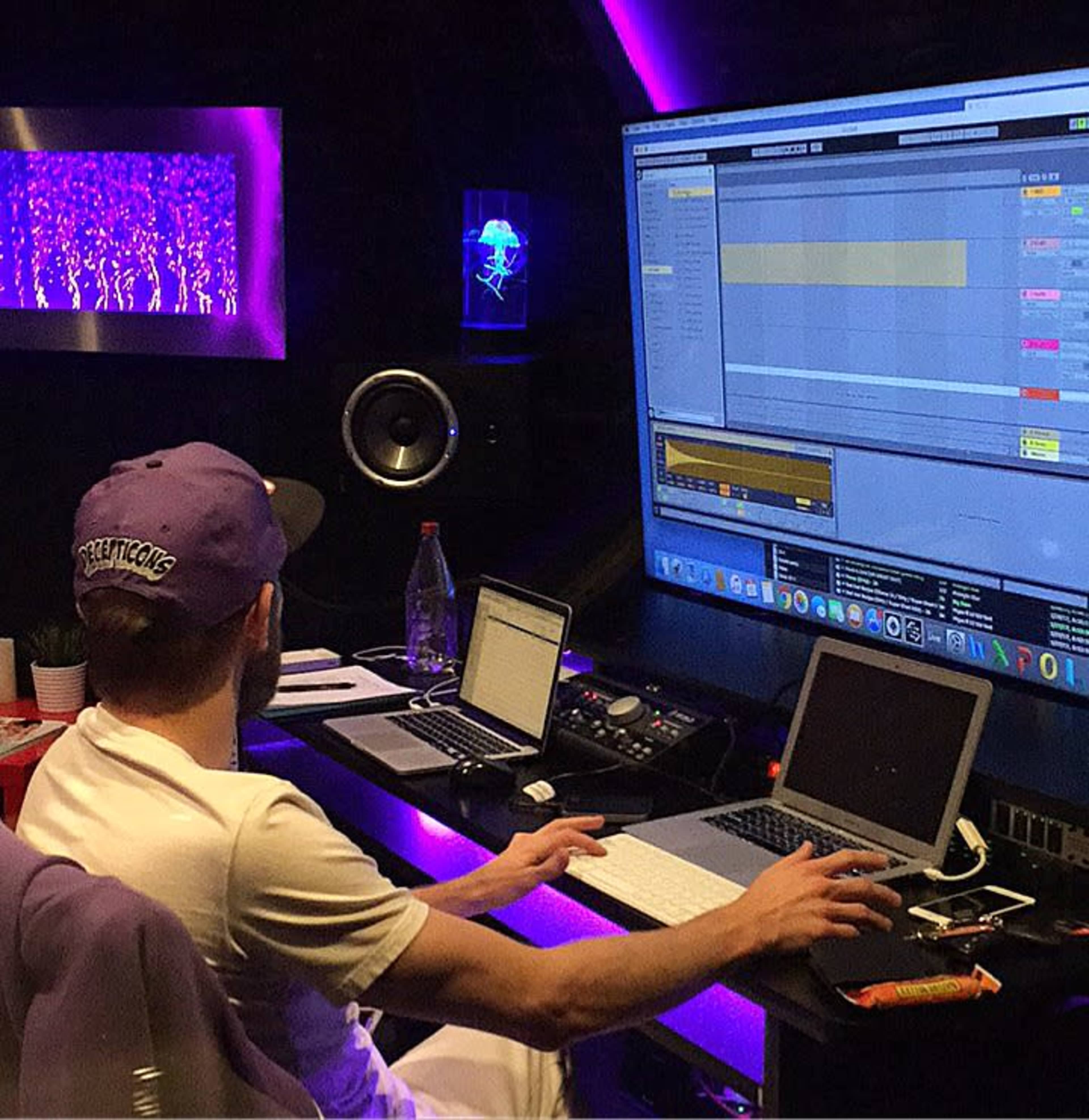 A person works on music production in a dimly lit studio with multiple computer screens and colorful lighting.