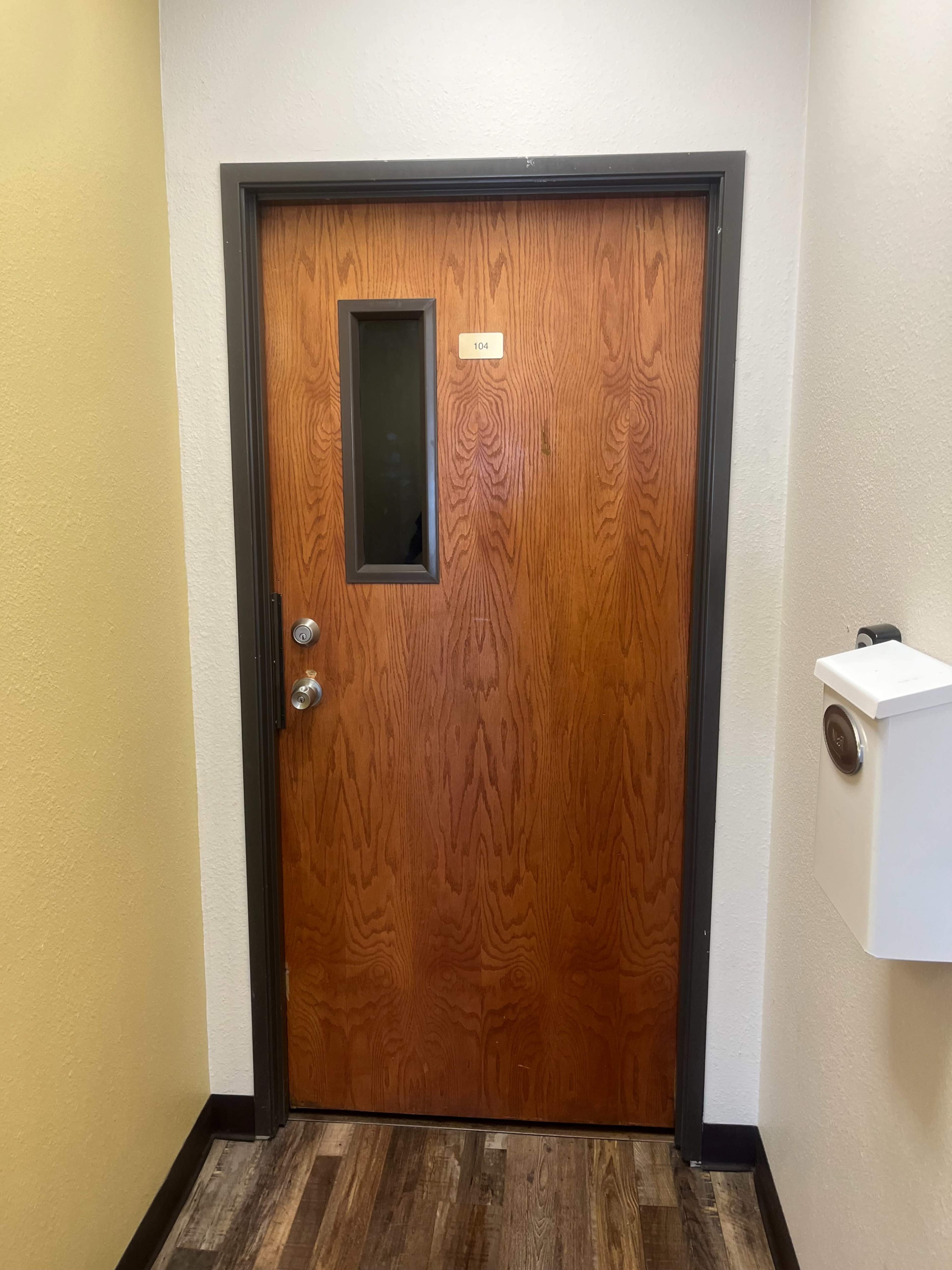 The image shows a wooden door with a small window, located in a brightly colored hallway with wooden flooring.