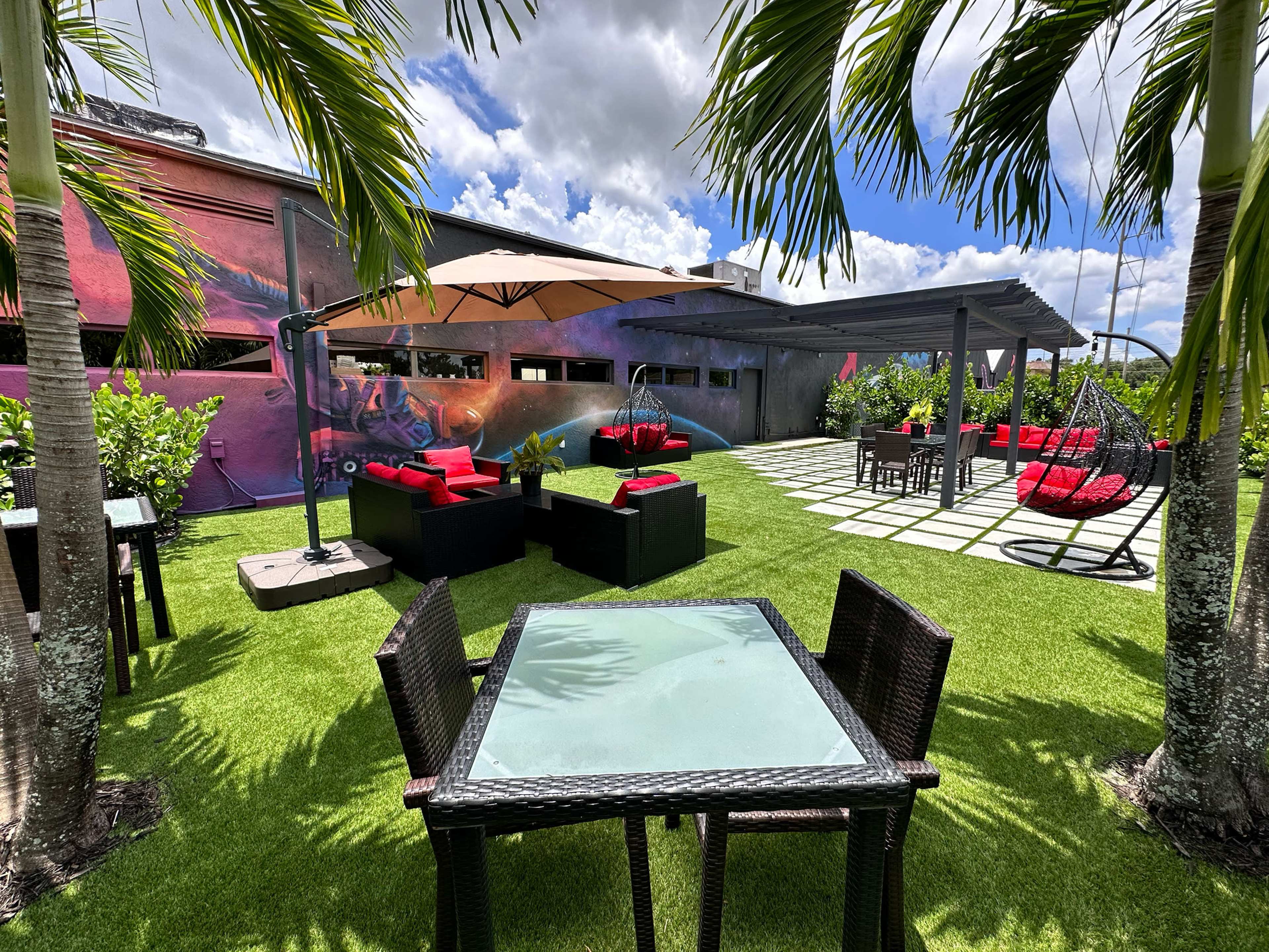 The image shows a rooftop garden area featuring tables and chairs, a shaded umbrella, and lounge seating surrounded by greenery and a colorful mural.