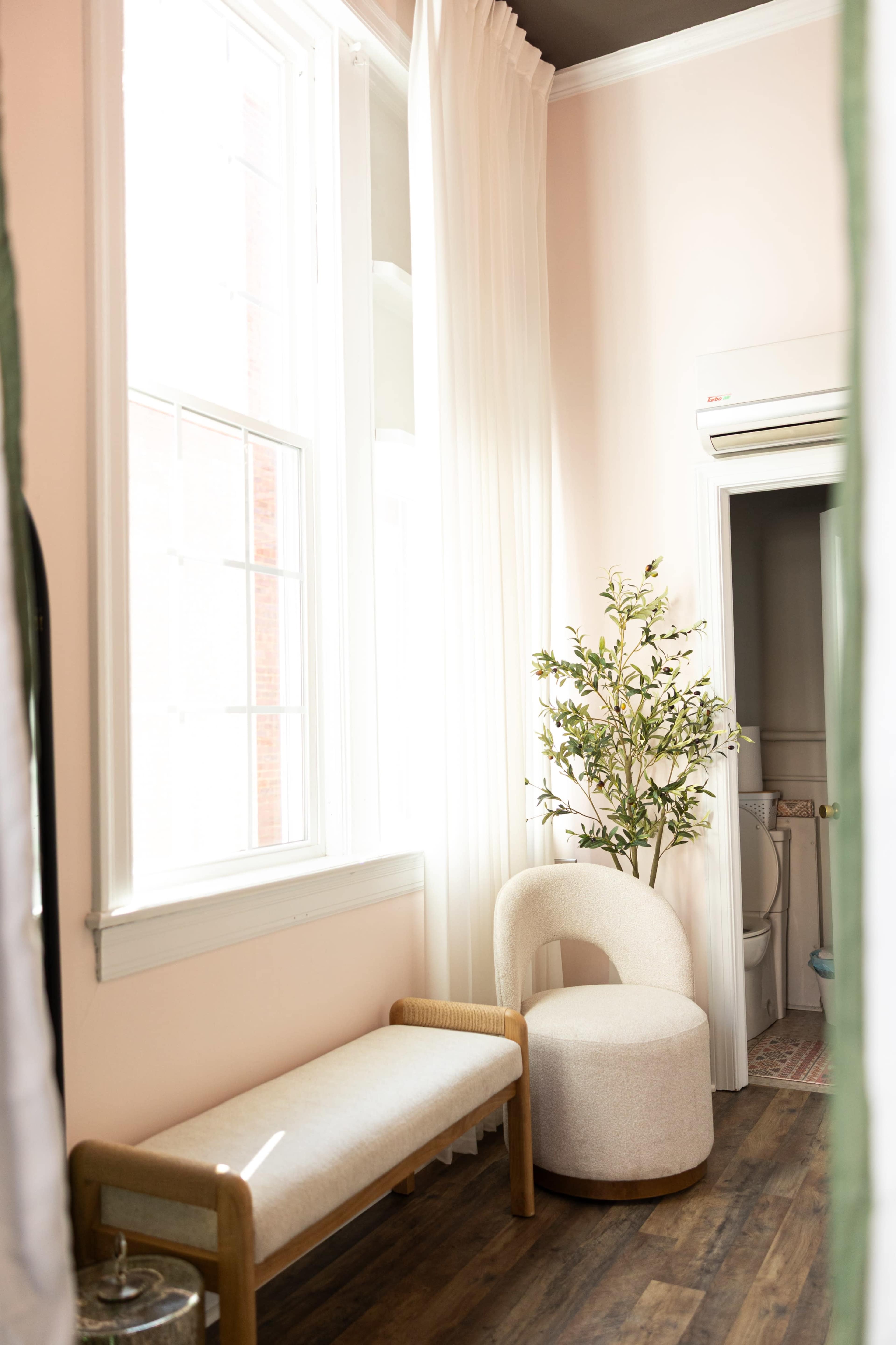 The image shows a brightly lit room with a beige bench, a round upholstered chair, and a potted plant near large windows with sheer white curtains.