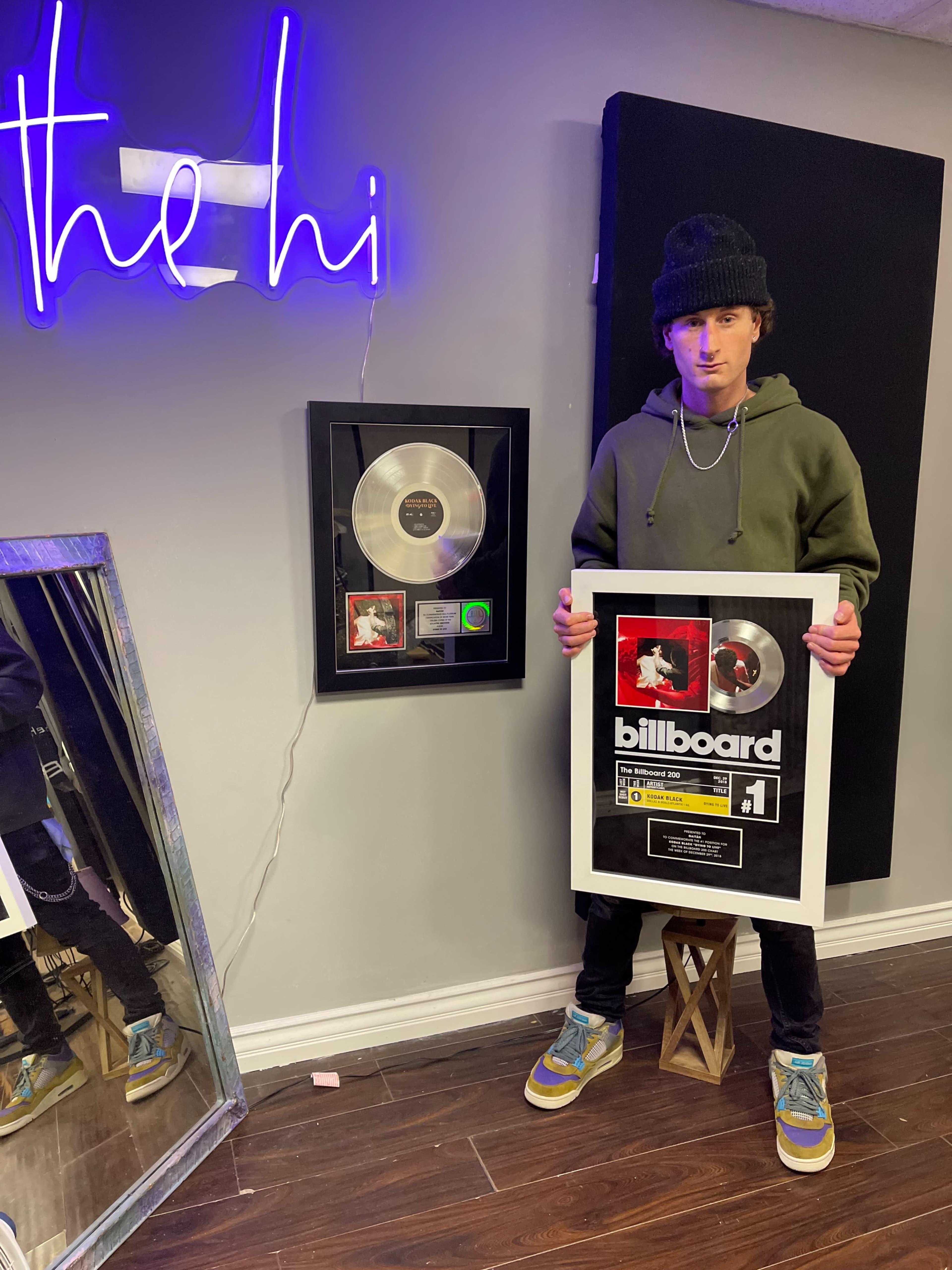 A person wearing a green hoodie and beanie stands next to a framed silver record plaque and a neon sign that says "the hi."