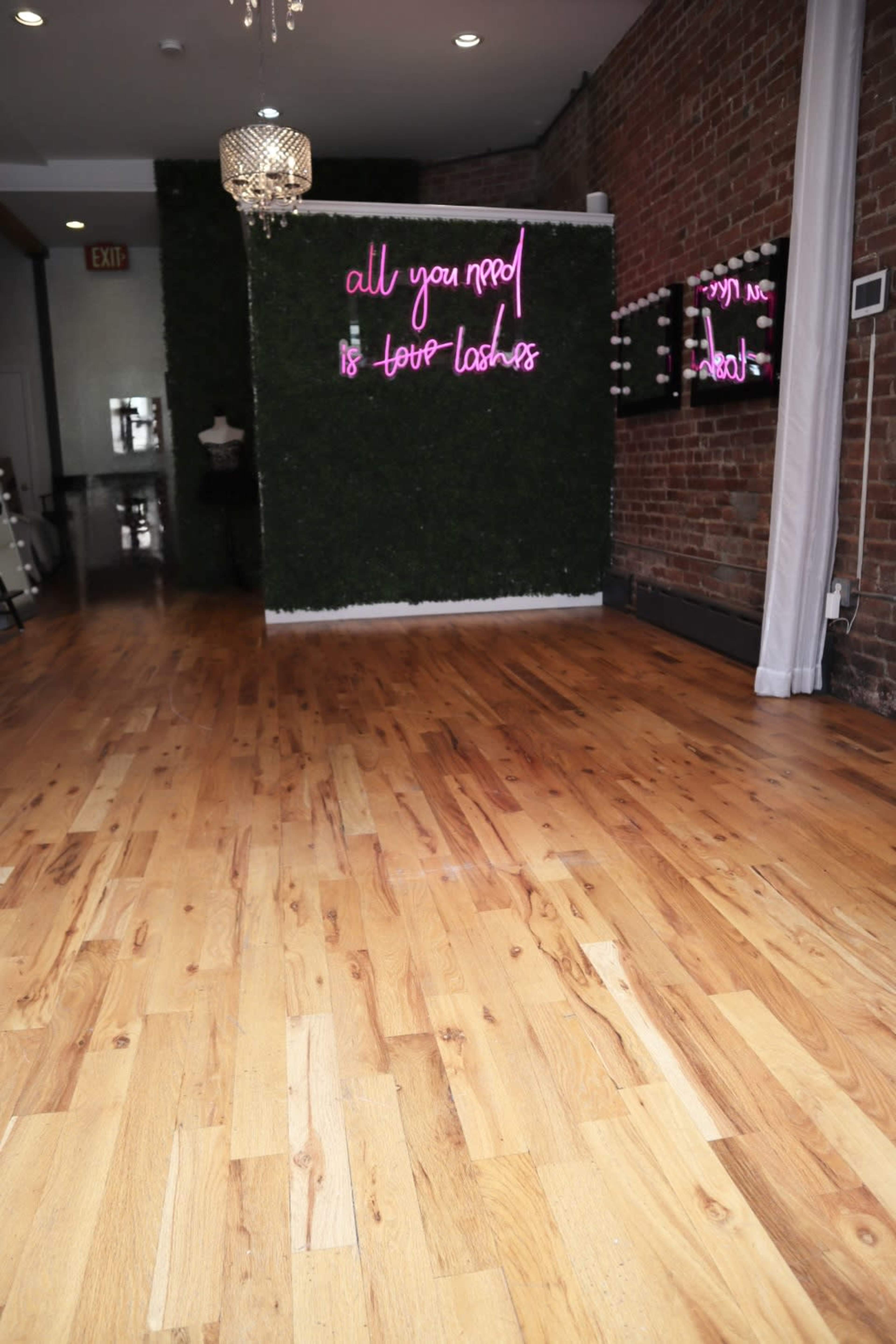 The image shows a room with wooden flooring, a greenery-covered wall featuring neon pink text, and a vanity area with mirrors and lights.