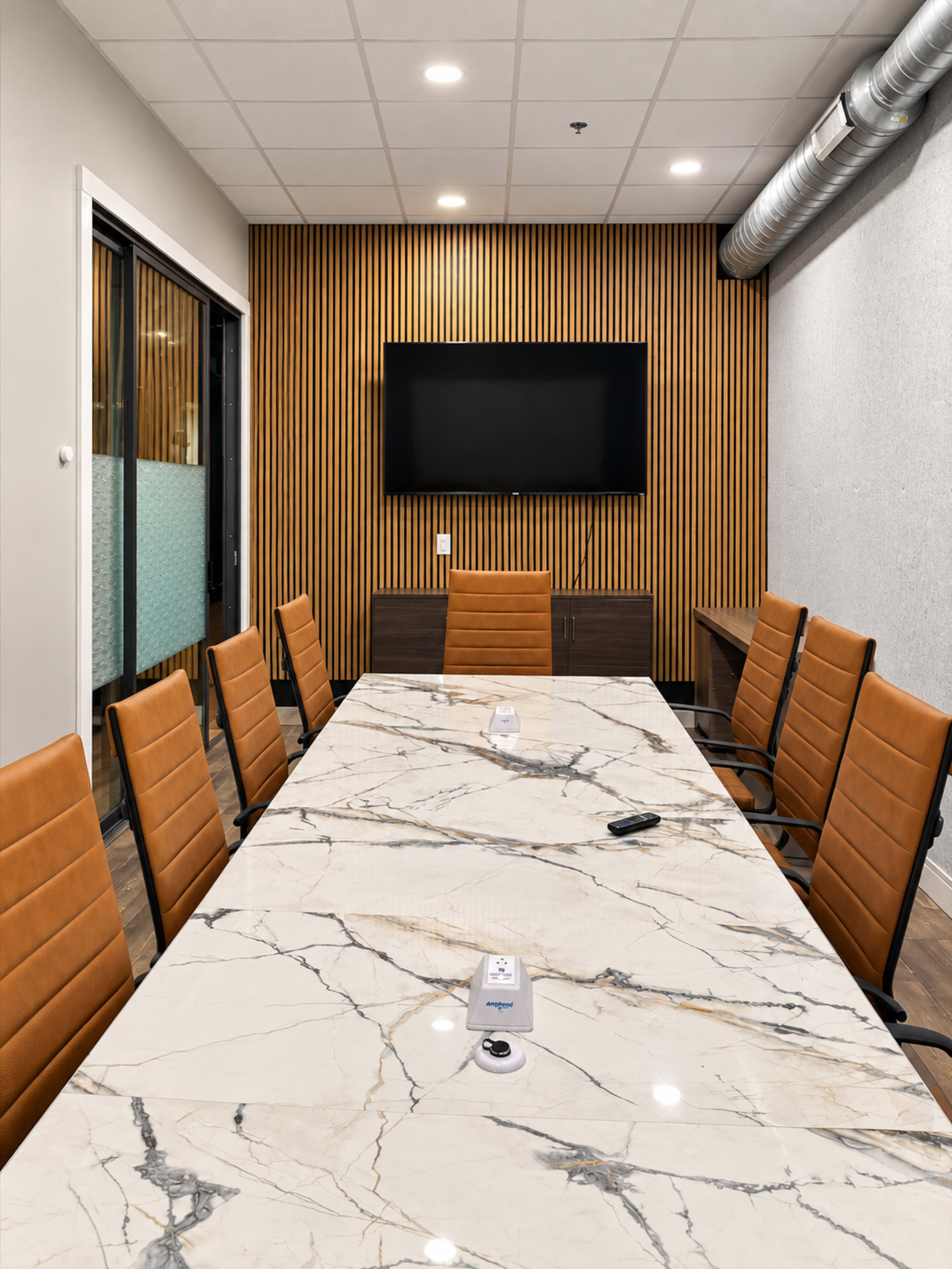The image shows a modern conference room with a large marble table, brown leather chairs, and a wooden accent wall behind a wall-mounted television.
