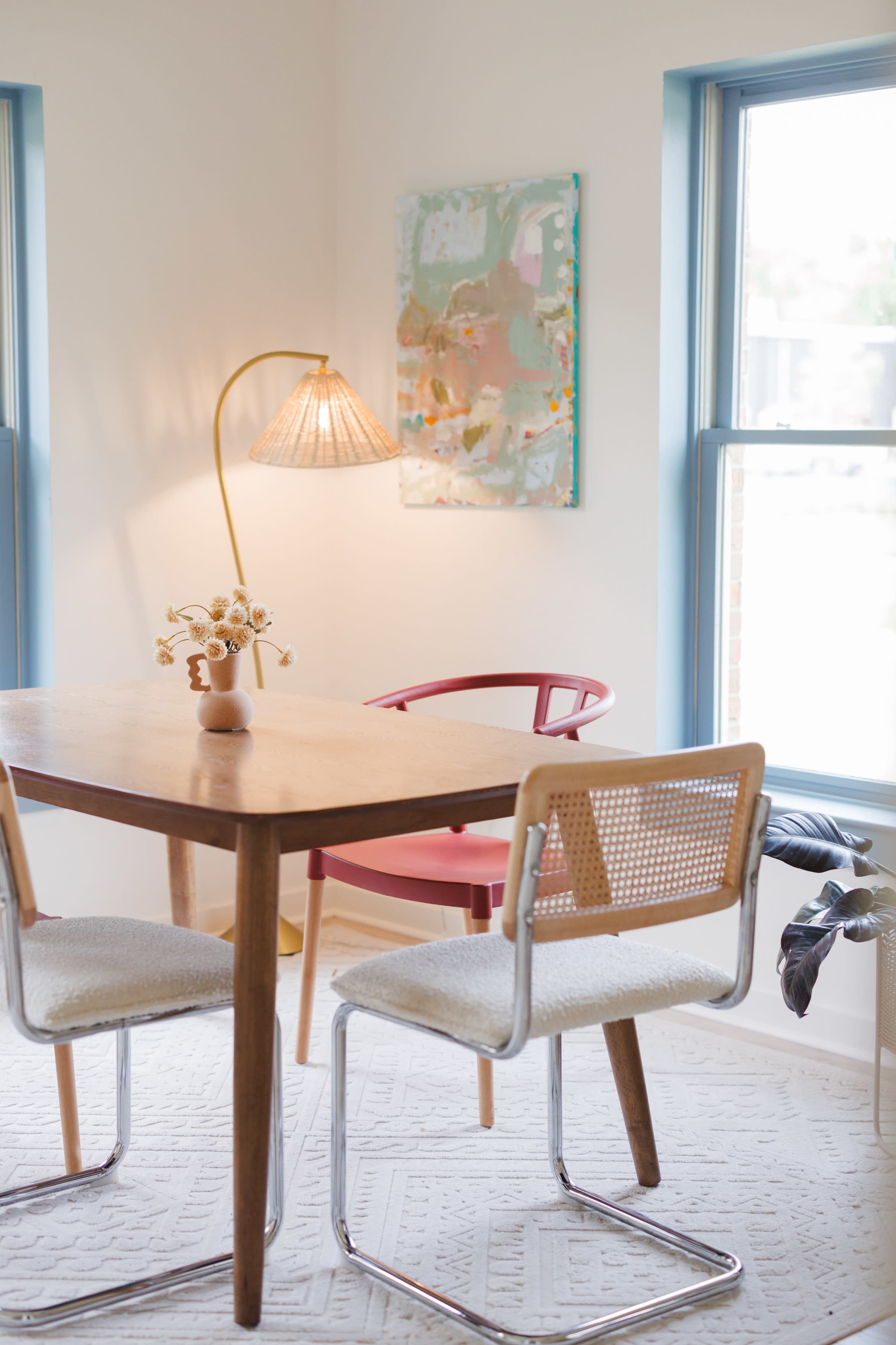 A wooden dining table with two chairs is positioned in a bright room featuring large windows, a floor lamp, and a colorful piece of artwork on the wall.