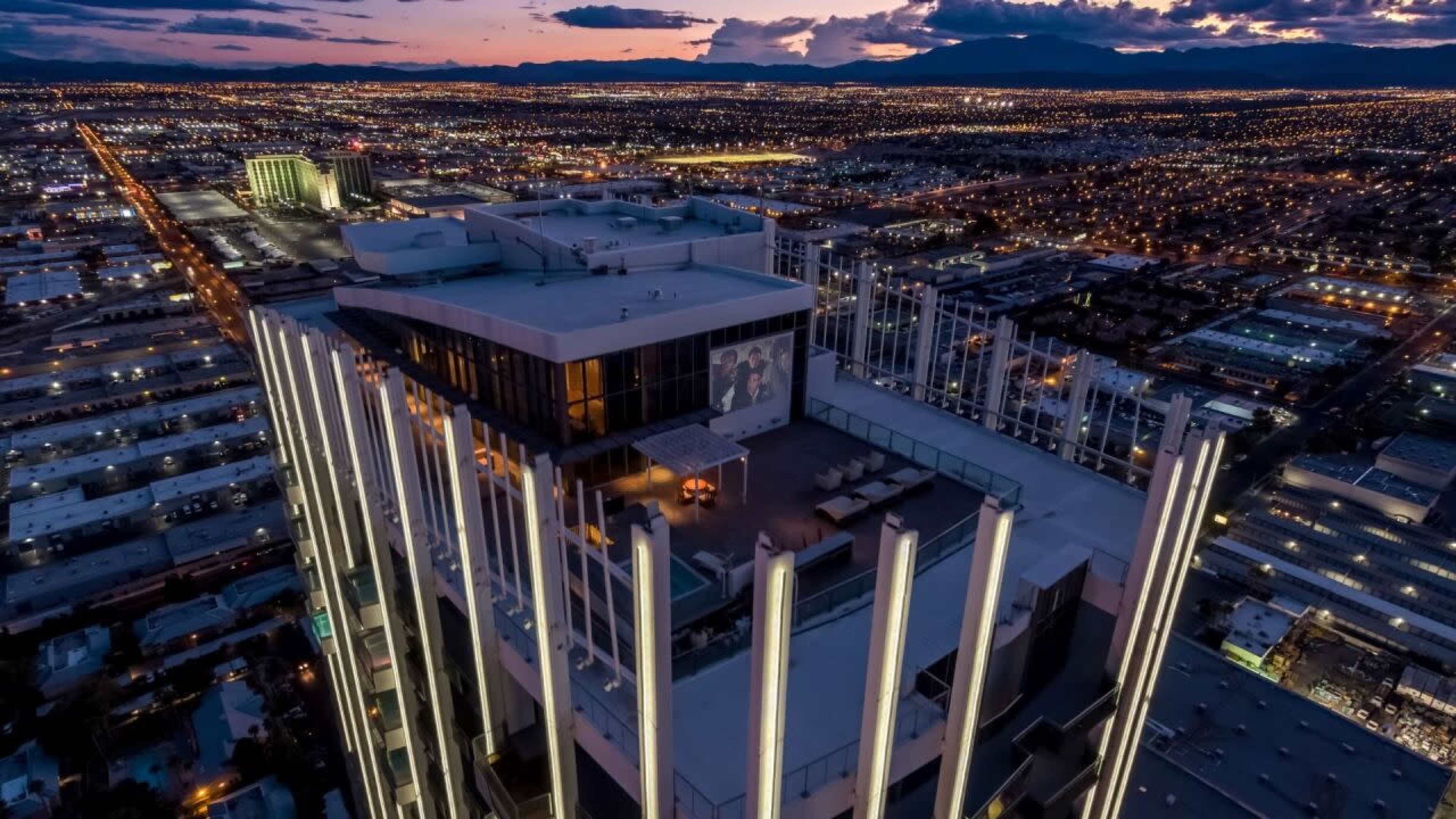 The image shows a high-rise building with illuminated vertical lights on its rooftop, overlooking a sprawling urban landscape at dusk.
