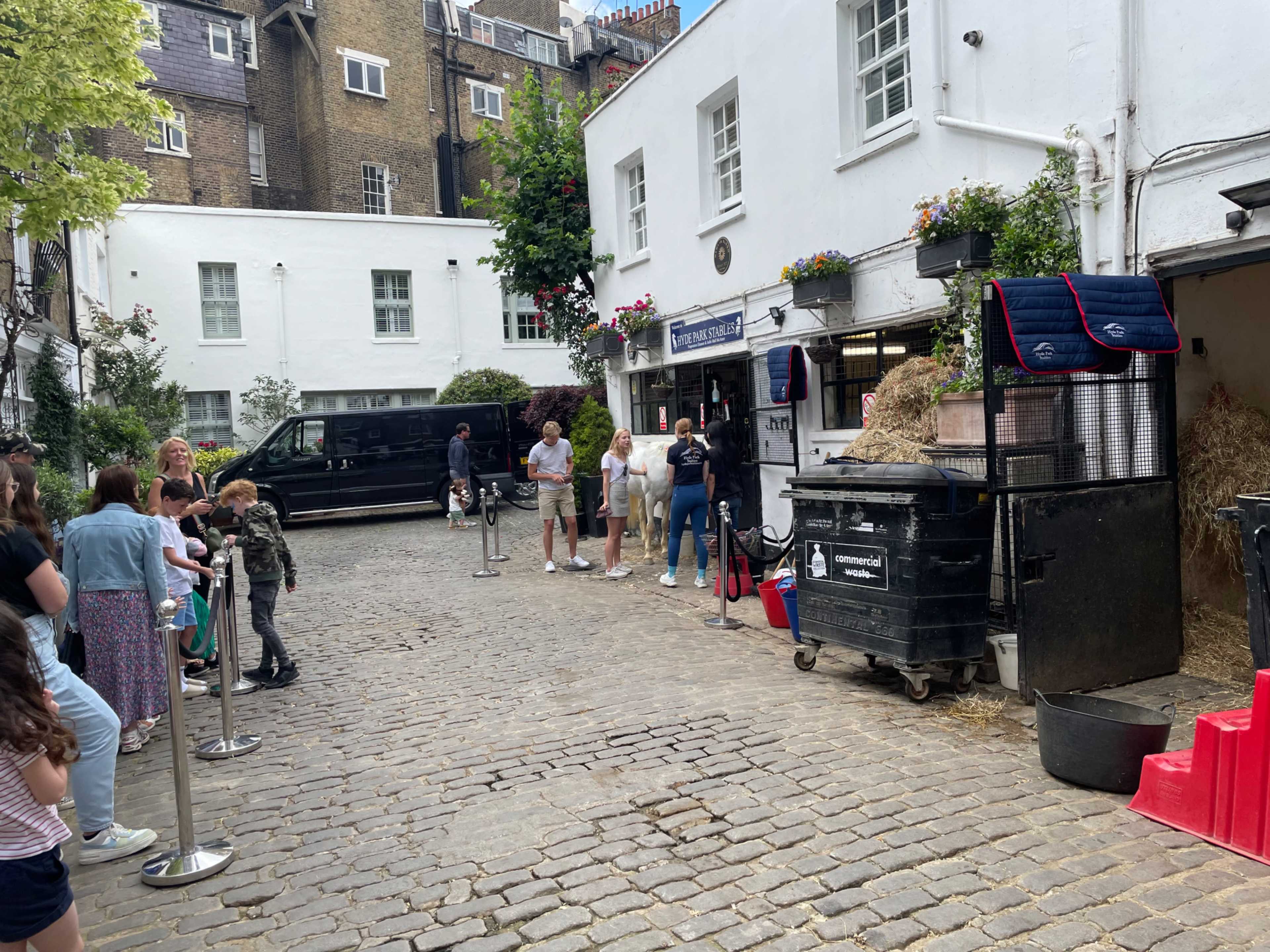 A crowd of people stands in line on a cobblestone street near a stable and trash bins, with a black vehicle parked in the background.