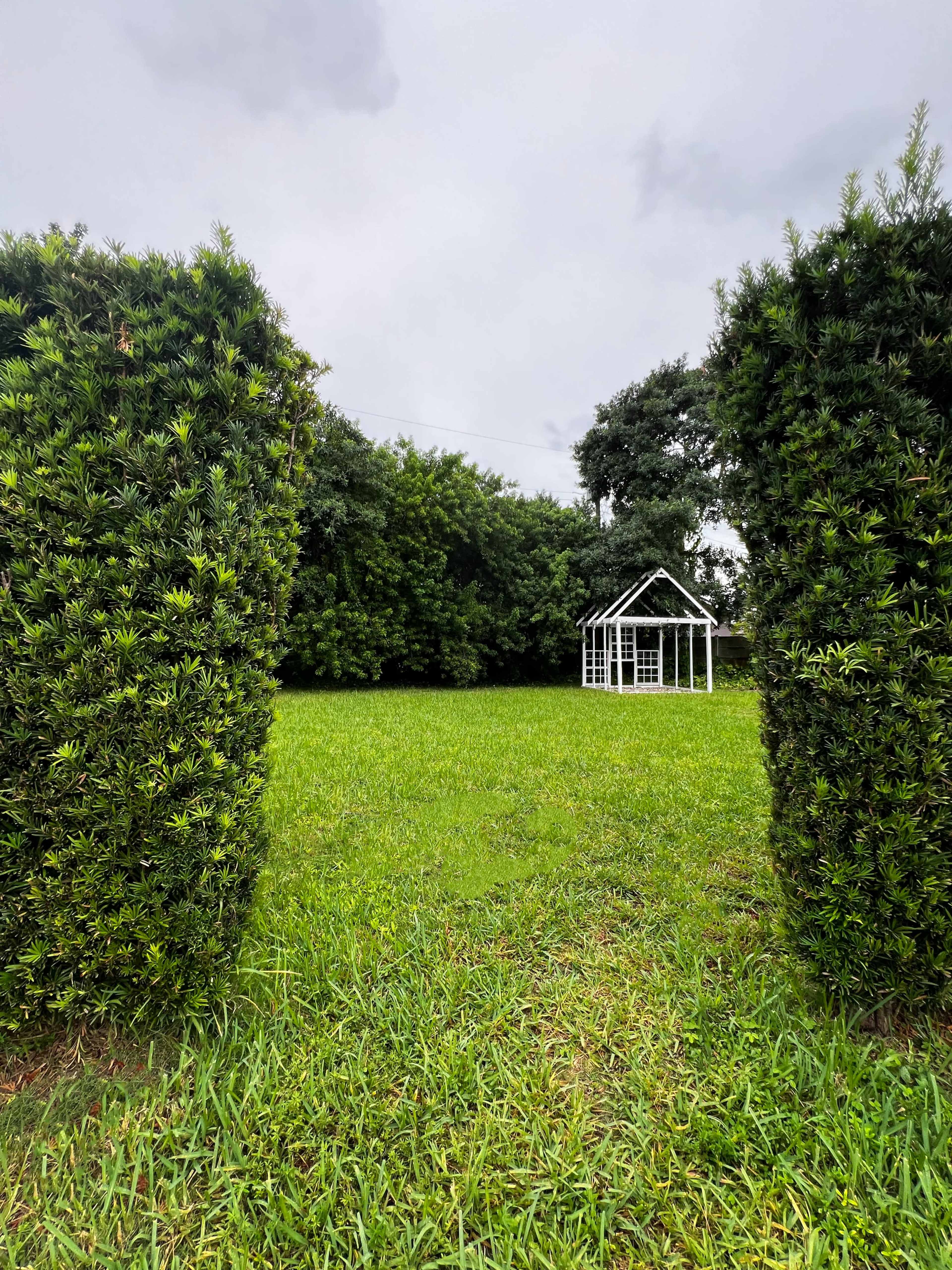 A green lawn is framed by tall hedges, leading to a small white structure in the distance.