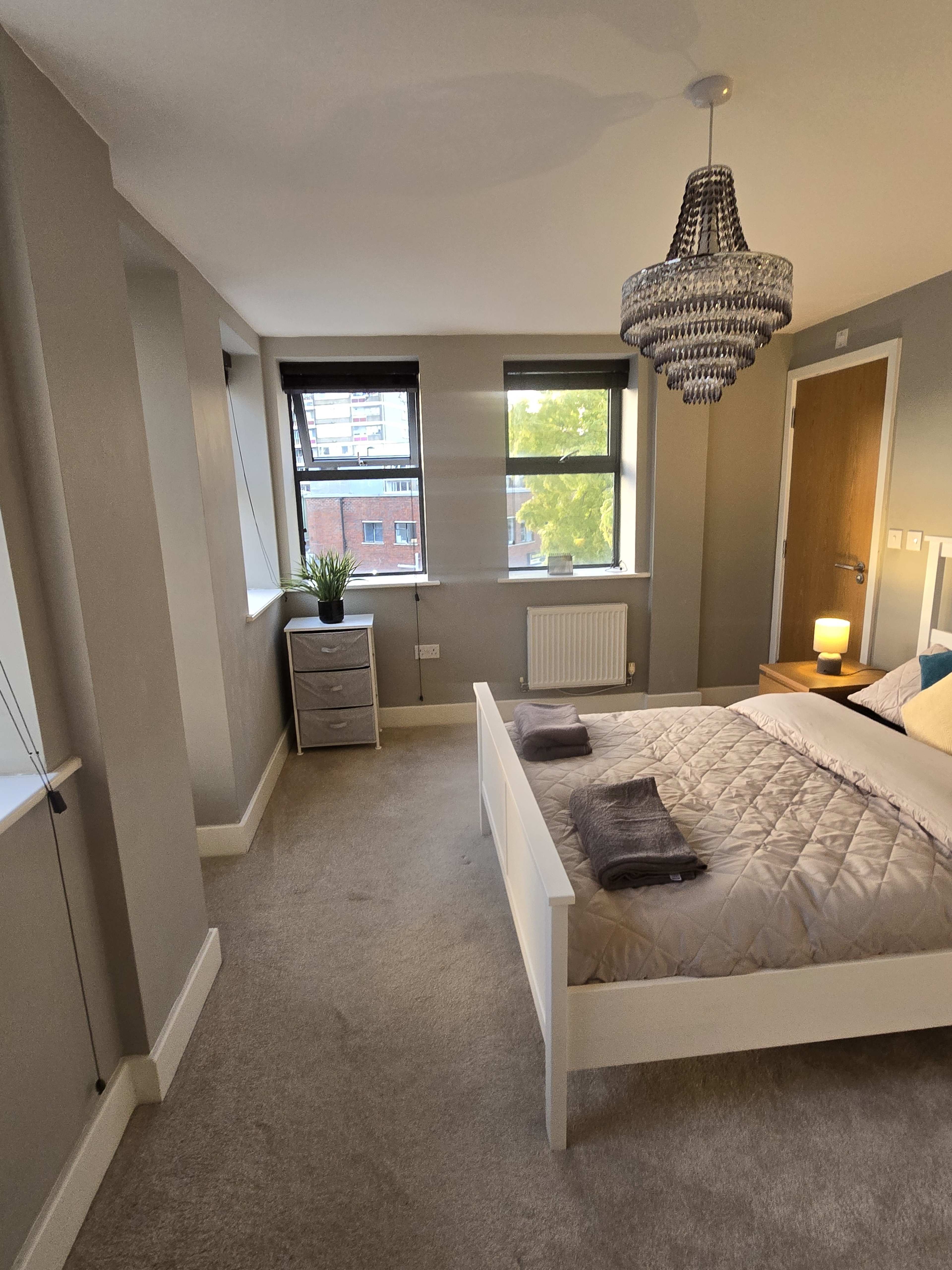 A neatly arranged bedroom features a bed with a quilt, a nightstand, and large windows that let in natural light.