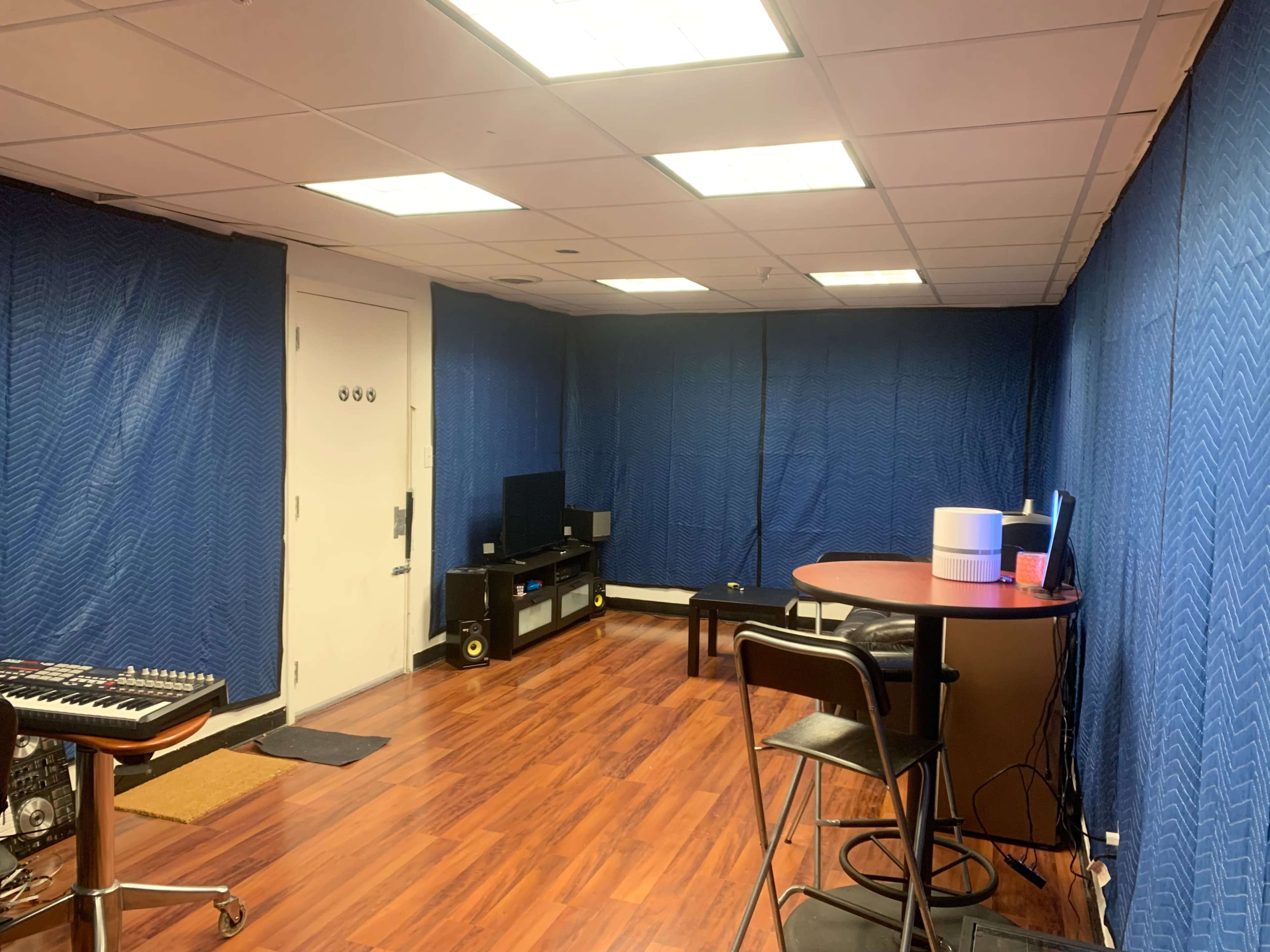 The room features blue sound-dampening curtains, a table with stools, a television, and a music keyboard against a wooden floor.