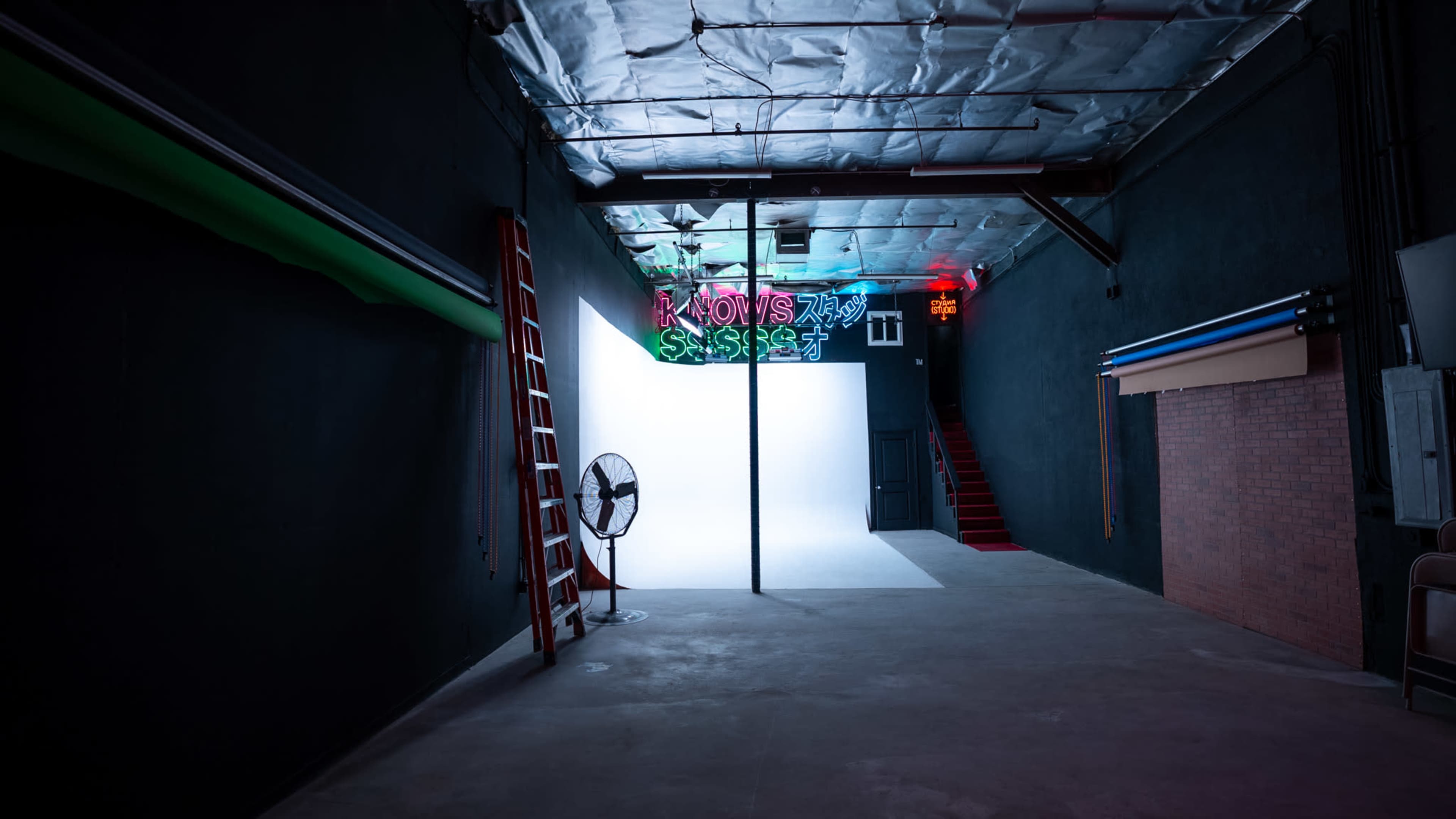 A spacious studio with a white backdrop, metal stairs, a fan, and colorful neon signs on the walls.