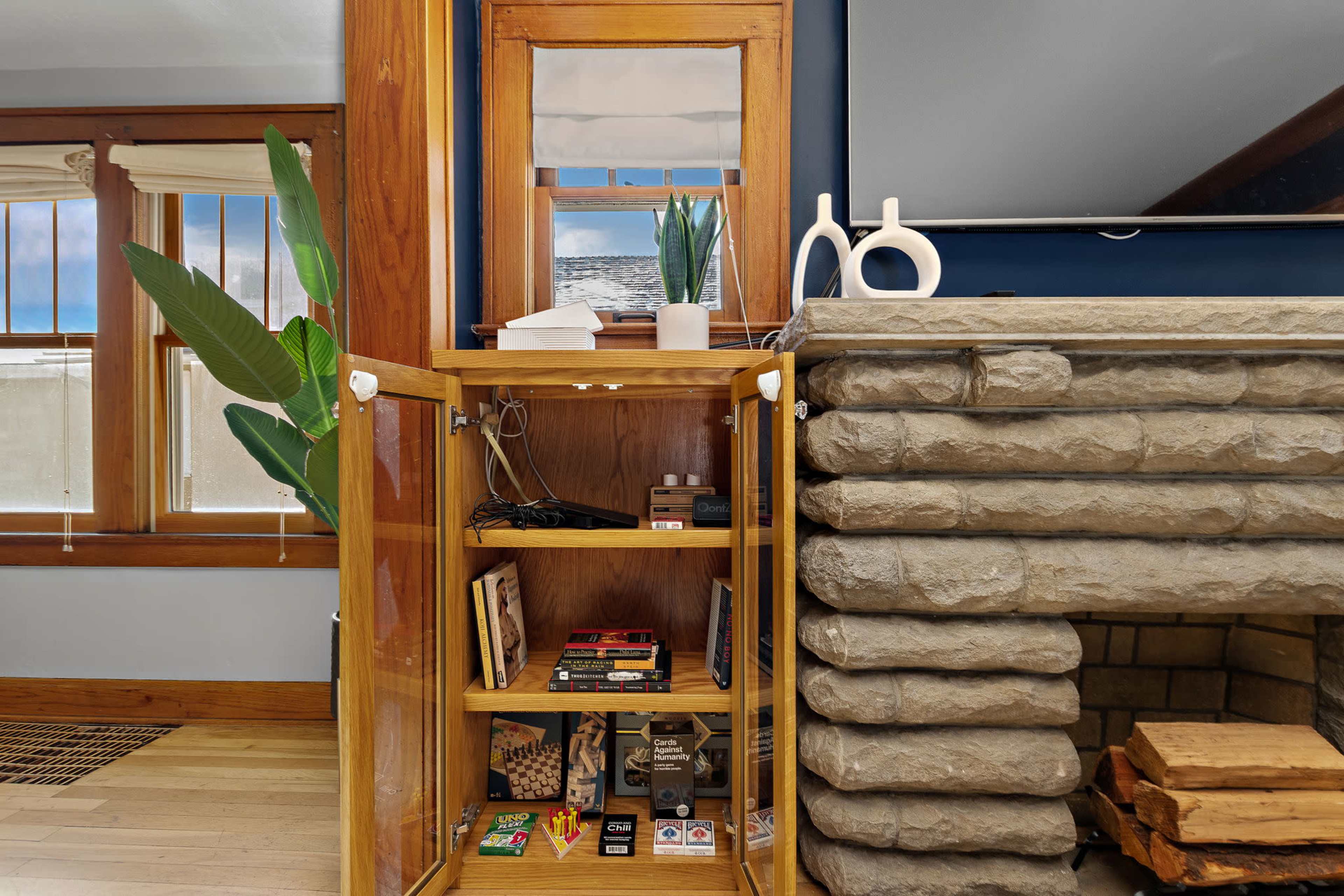 A wooden cabinet beside a stone fireplace displays various board games and supplies, while a plant and modern decor accent the room.