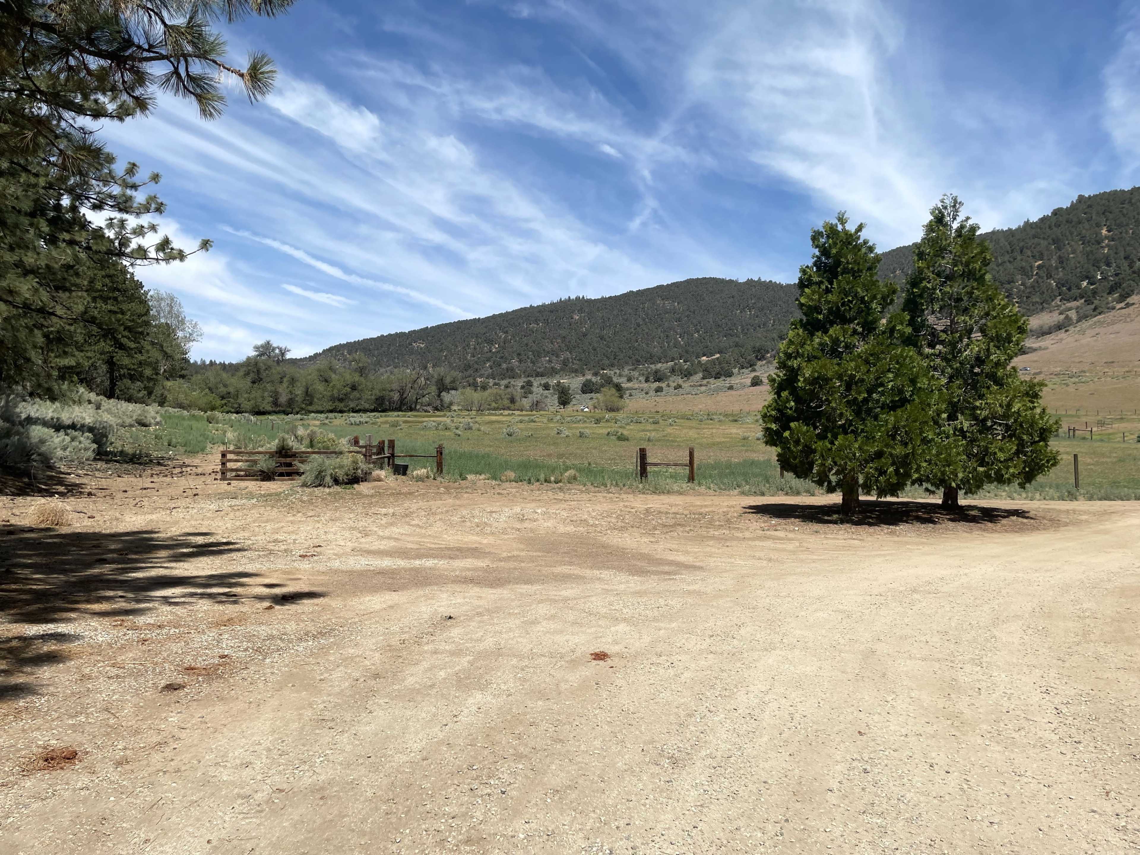 A dirt road leading to an open field surrounded by mountains and trees under a partly cloudy sky.