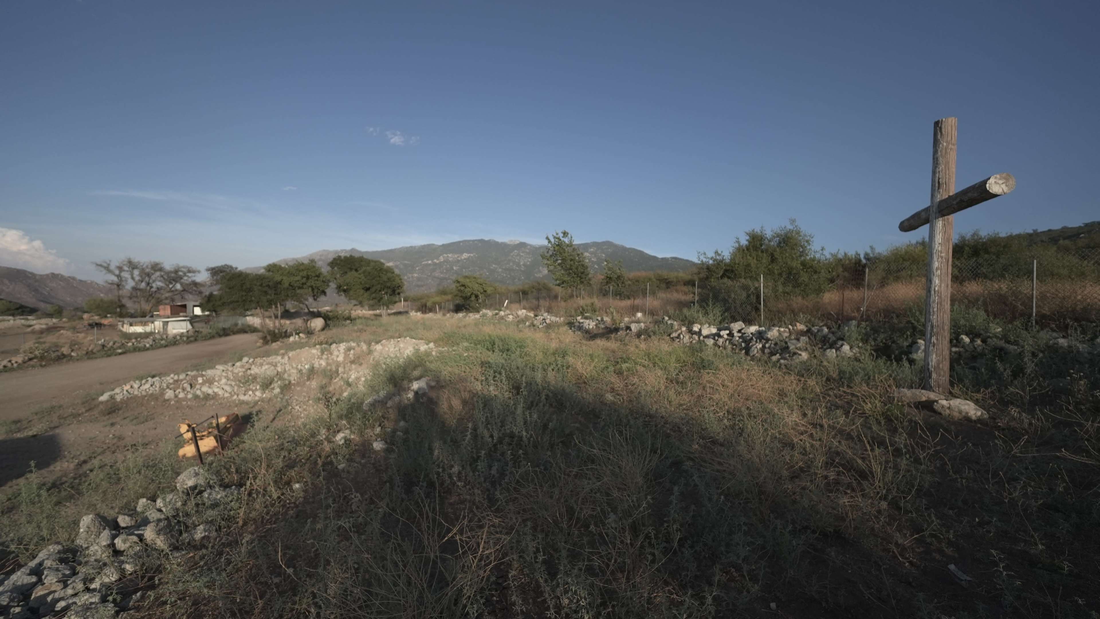The image shows a rural landscape with a wooden cross standing in a sandy area surrounded by sparse vegetation and rocky terrain.