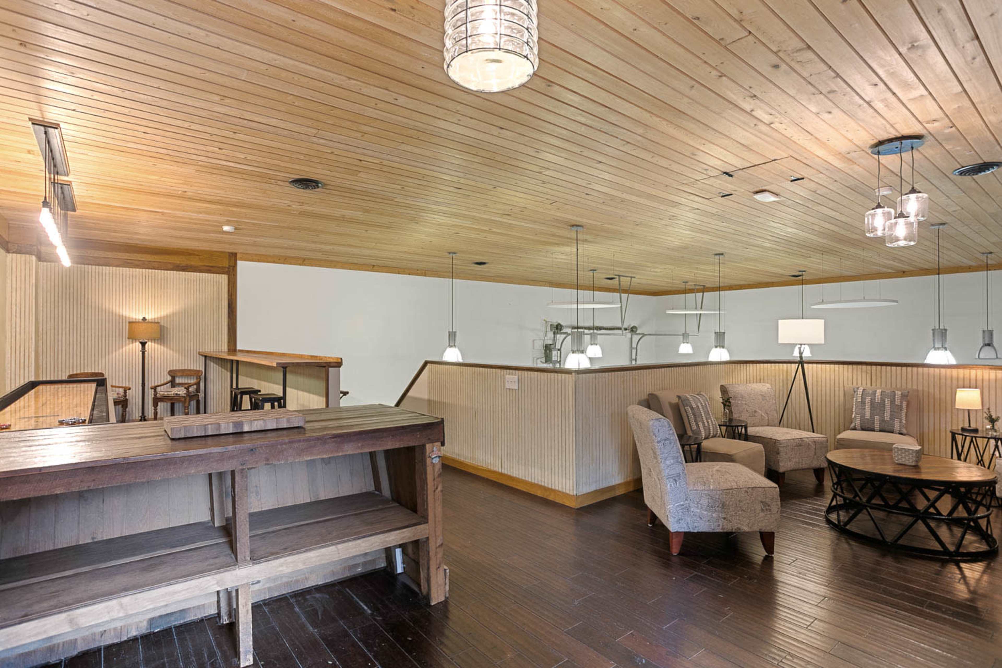 The image shows a spacious interior with wooden ceilings, a bar area, and seating arrangements that include a round table and comfortable chairs.