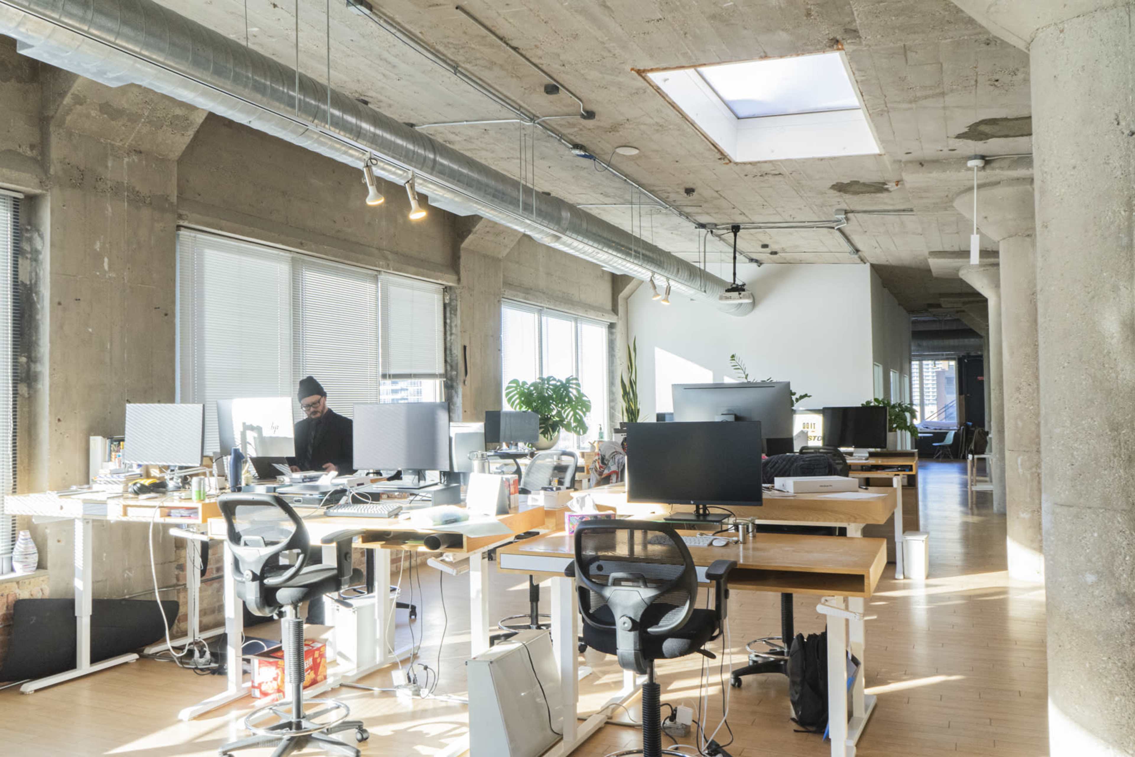 A spacious office with several workstations, large windows letting in natural light, and a person seated at a desk in the background.