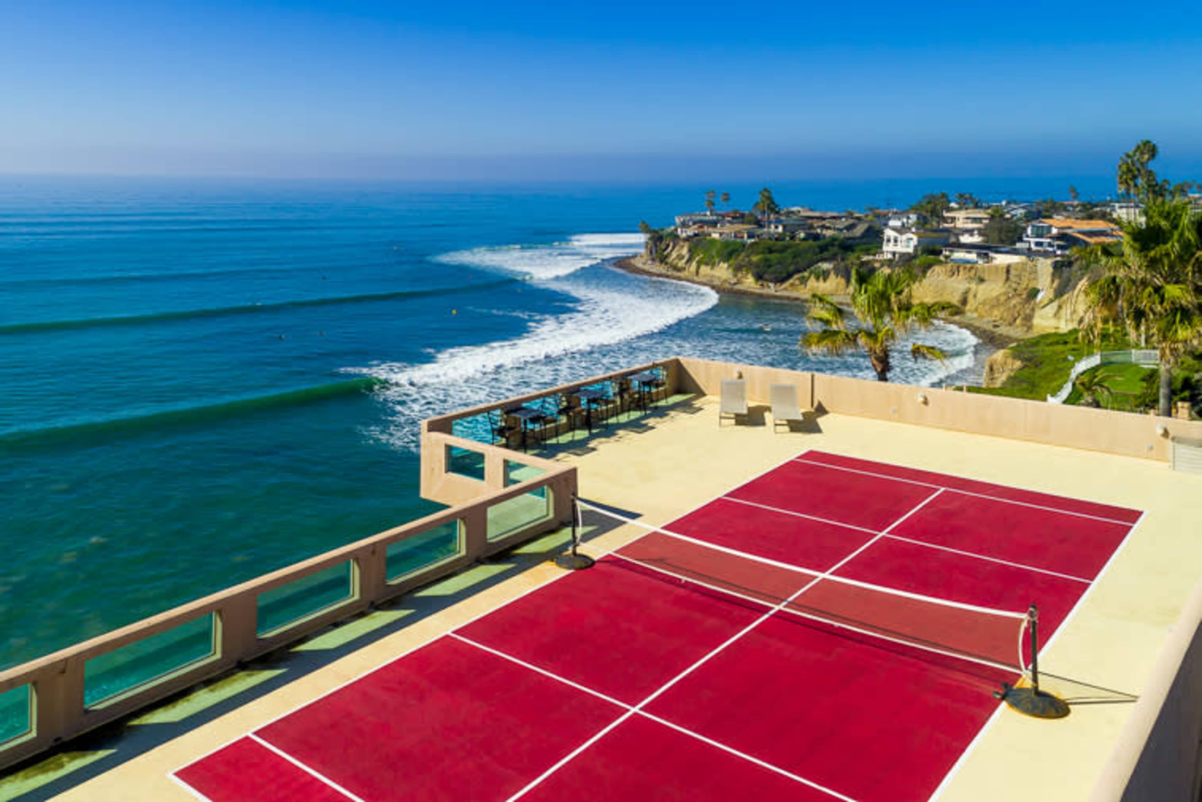 A seaside property with a red sports court overlooking a beach and ocean waves.