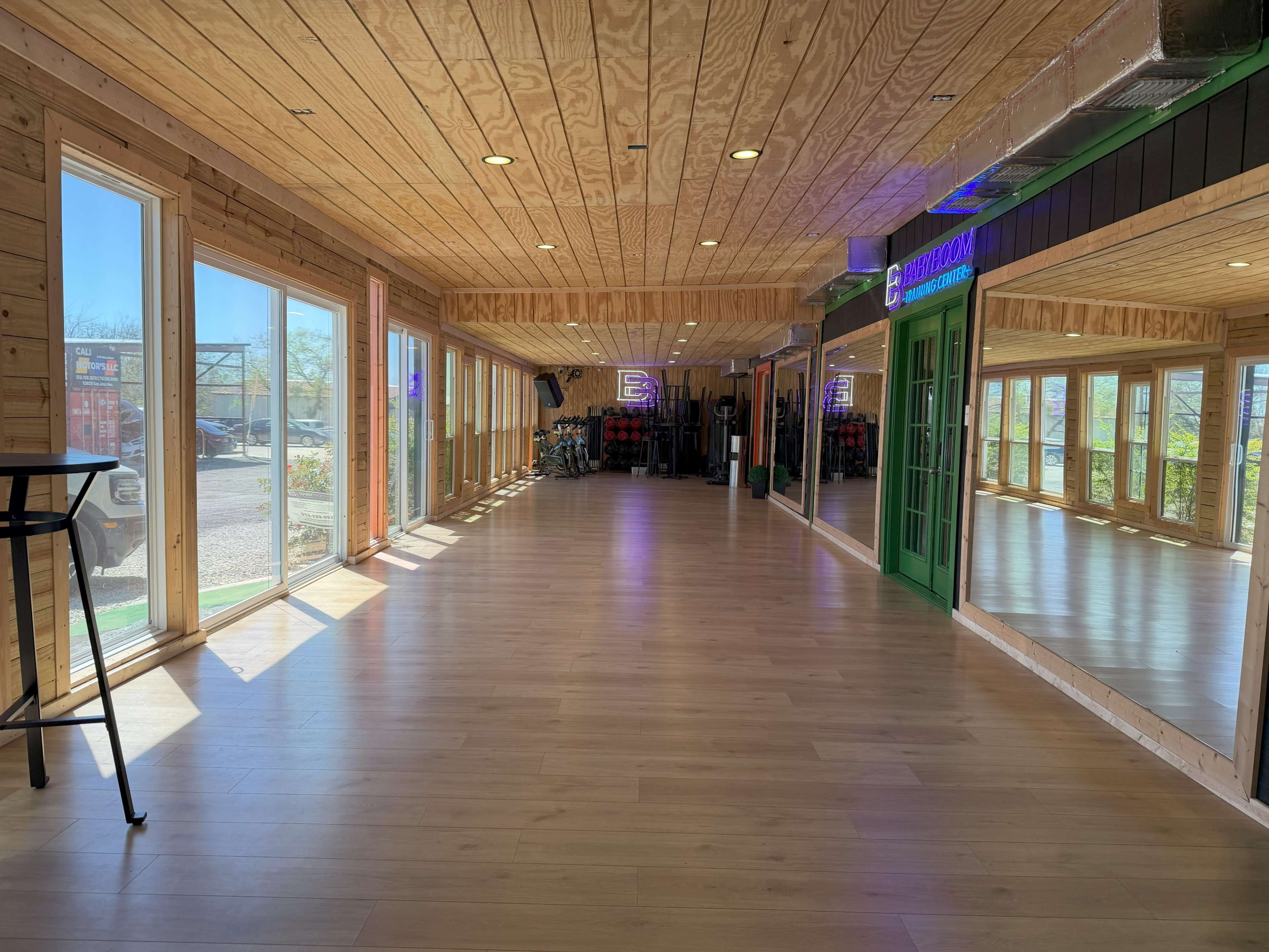 The image shows a spacious, wooden-floored room with large windows providing natural light and green doors leading to another area.