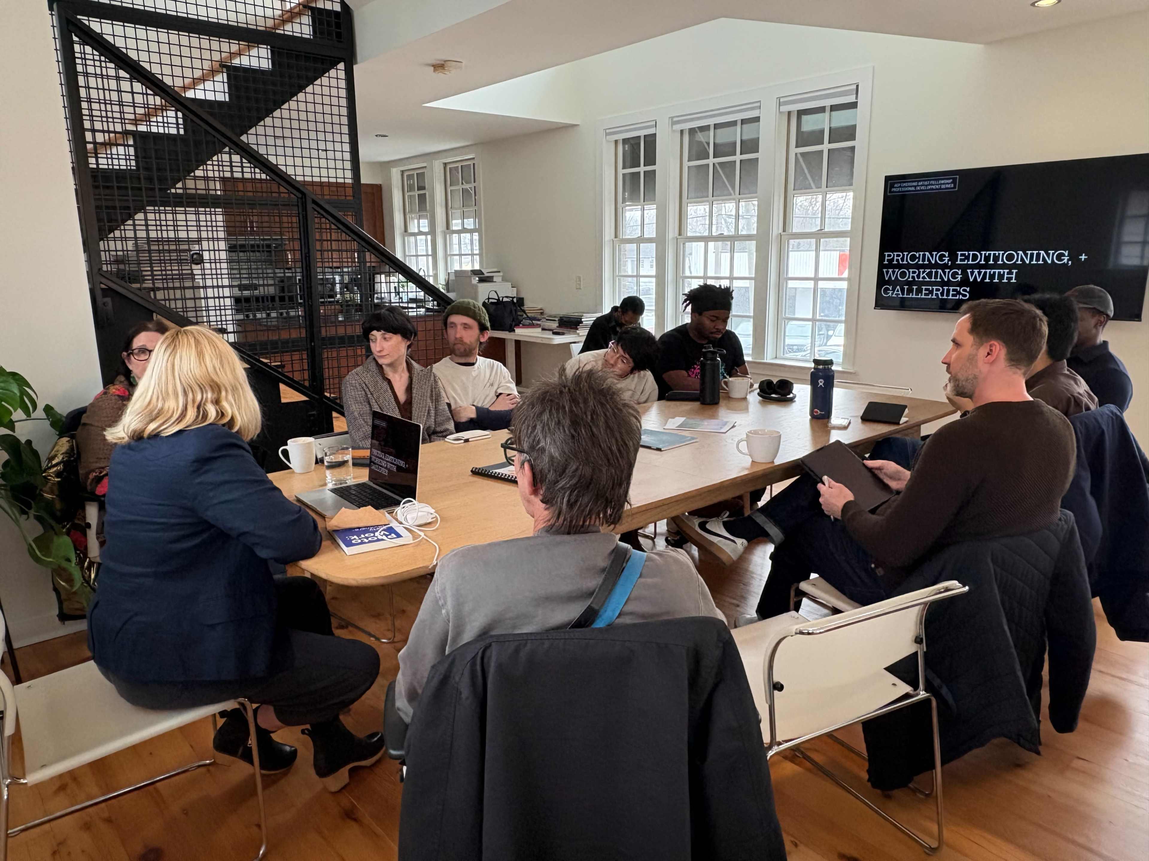 A group of people is seated around a table in a modern meeting space, engaged in a discussion about pricing, editioning, and working with galleries.