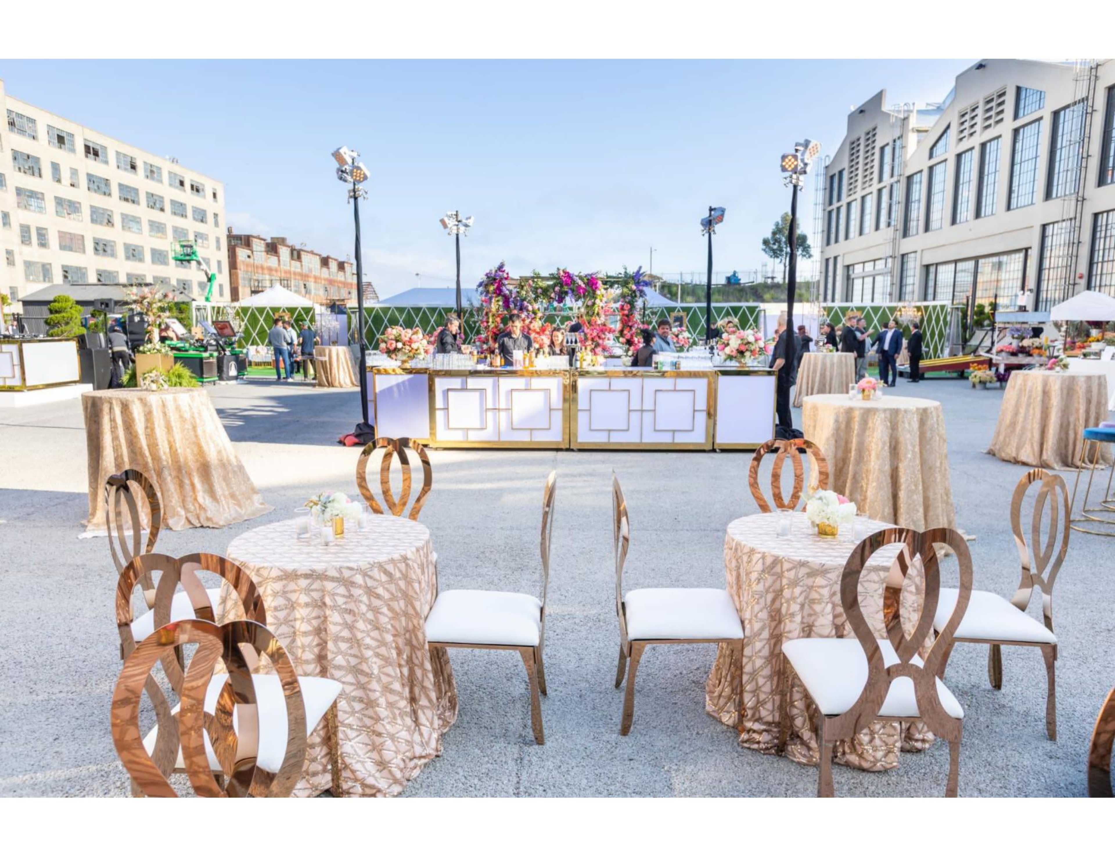 An outdoor event setup with round tables adorned in gold tablecloths and a decorated buffet area in the background.