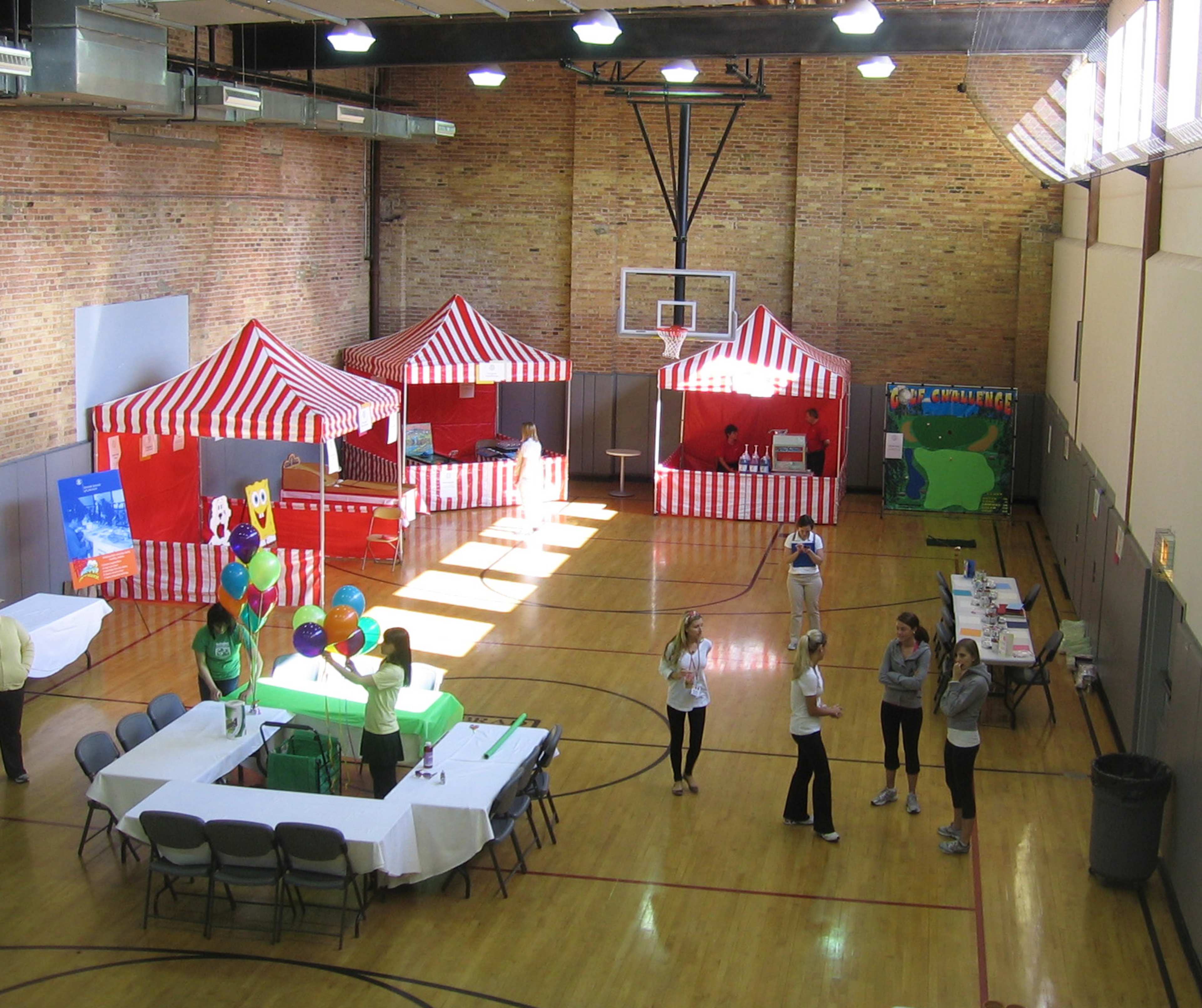 The image shows a gymnasium set up for an event, featuring two red and white striped tents, tables, and a basketball hoop in the background.