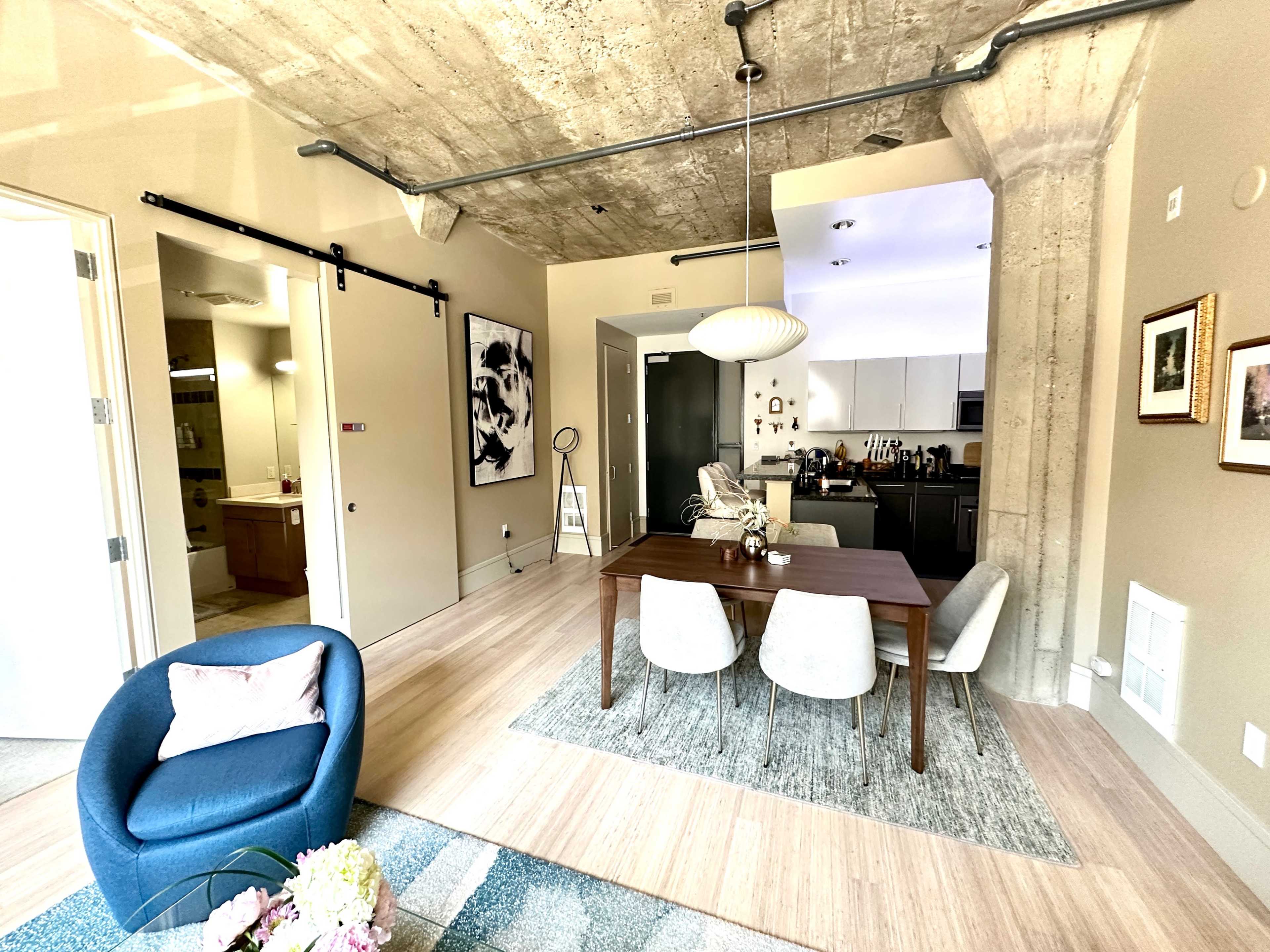 A modern living space featuring a dining area with a wooden table and white chairs, a blue armchair, and a kitchen visible in the background.