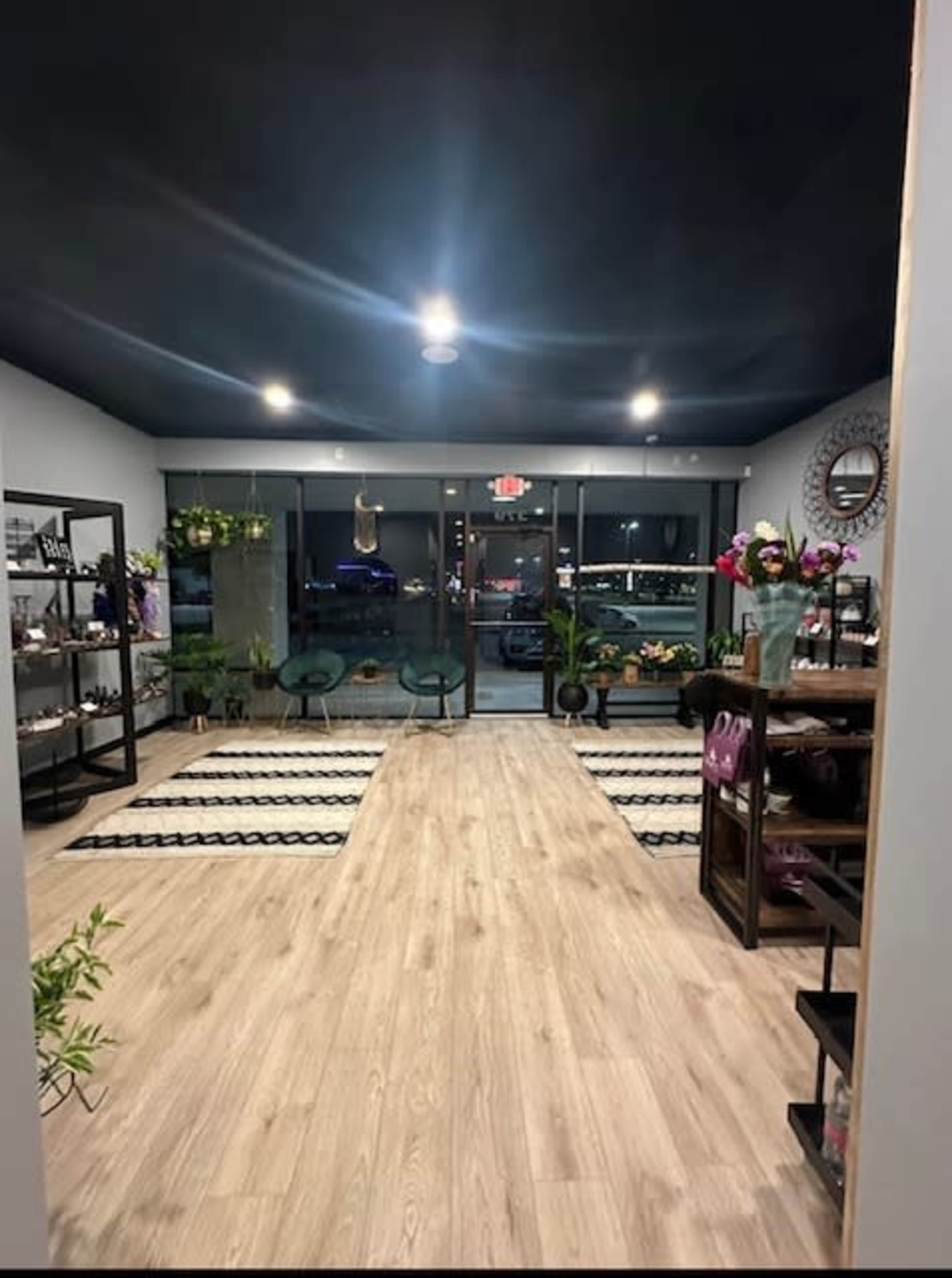 The image shows a modern interior of a shop with wooden flooring, plants, and seating areas, illuminated by overhead lights.