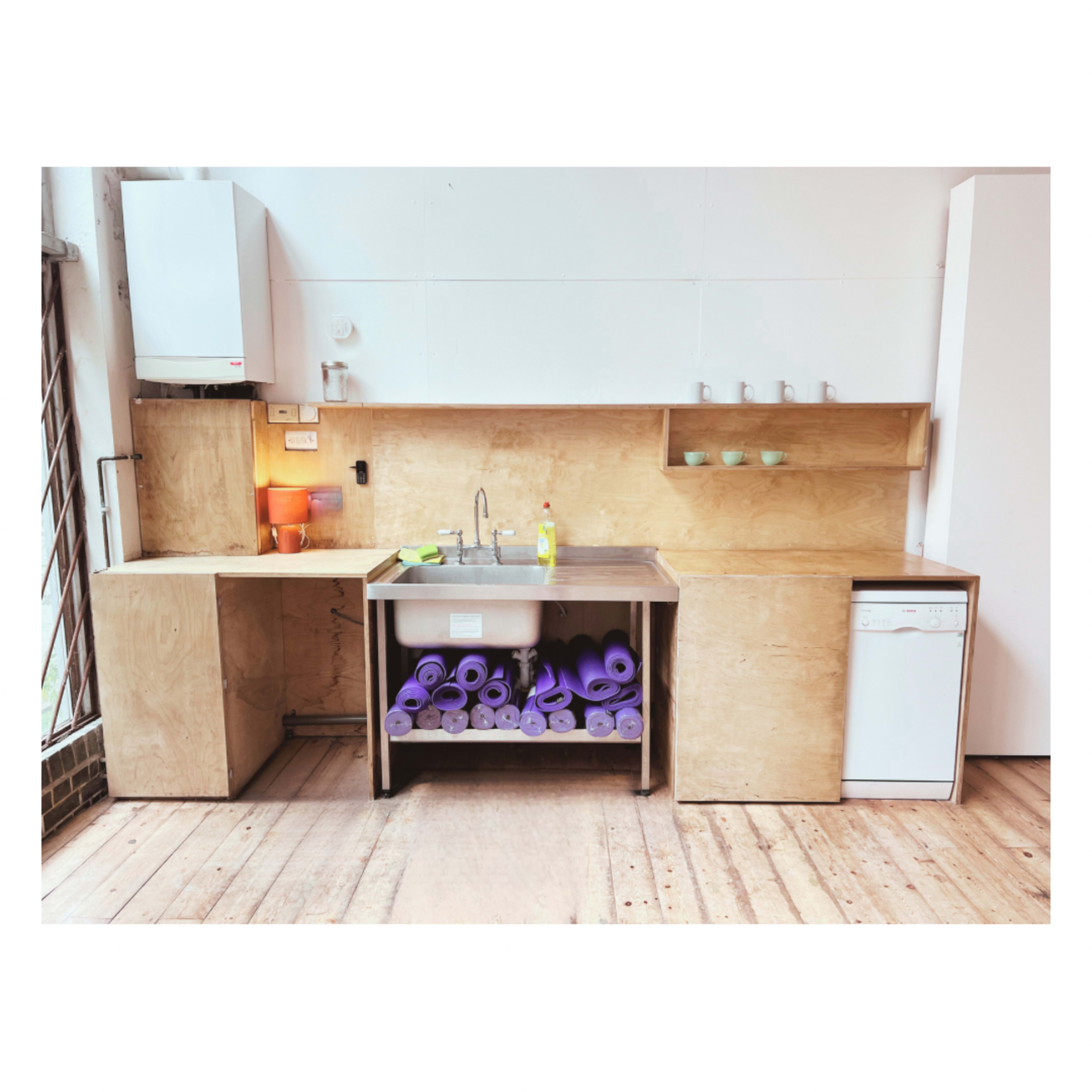 A minimalist kitchen with a stainless steel sink, a wooden countertop, and a stack of purple yoga mats stored underneath.