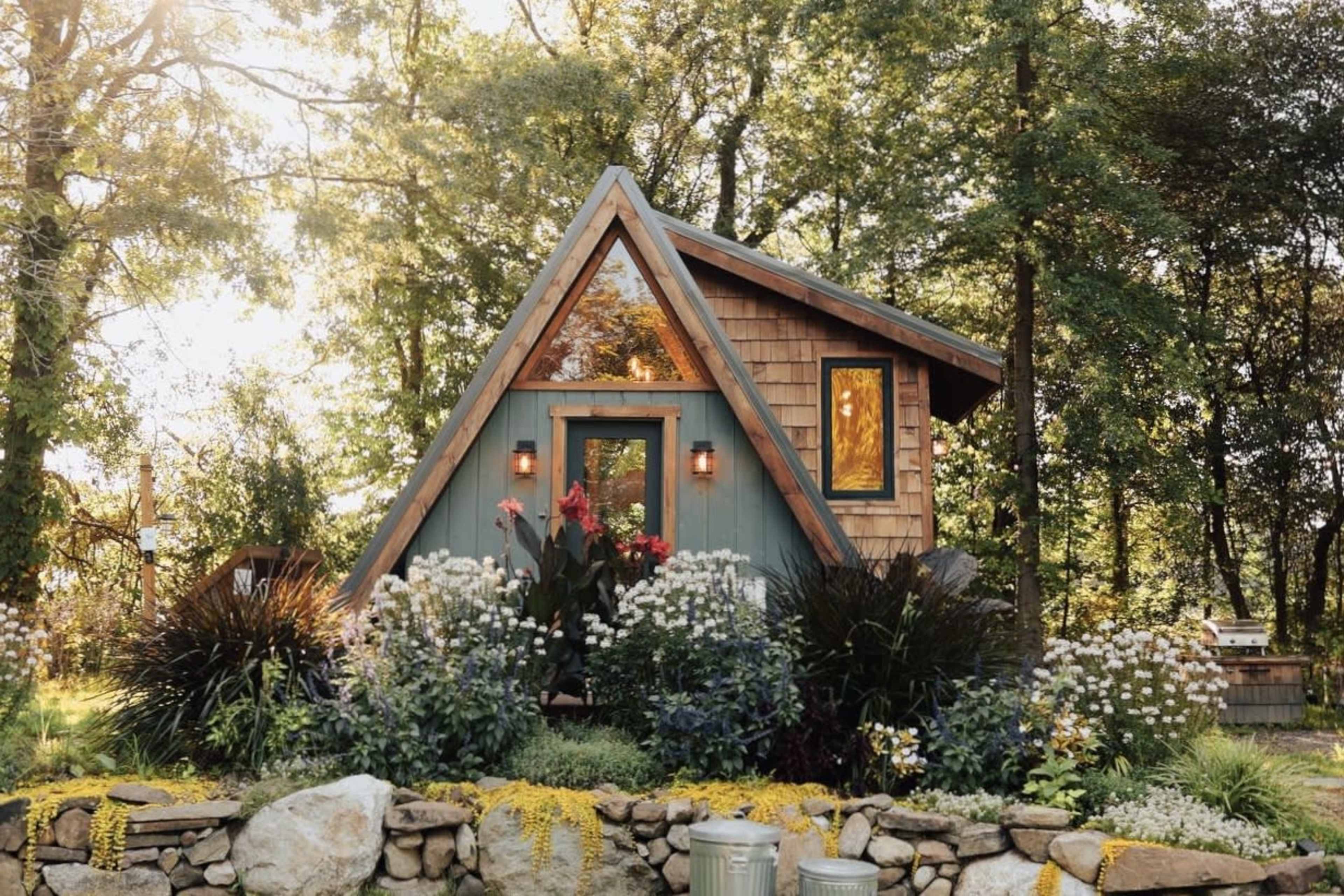 A small A-frame cabin nestled among trees, surrounded by a landscaped garden with various flowers and plants.
