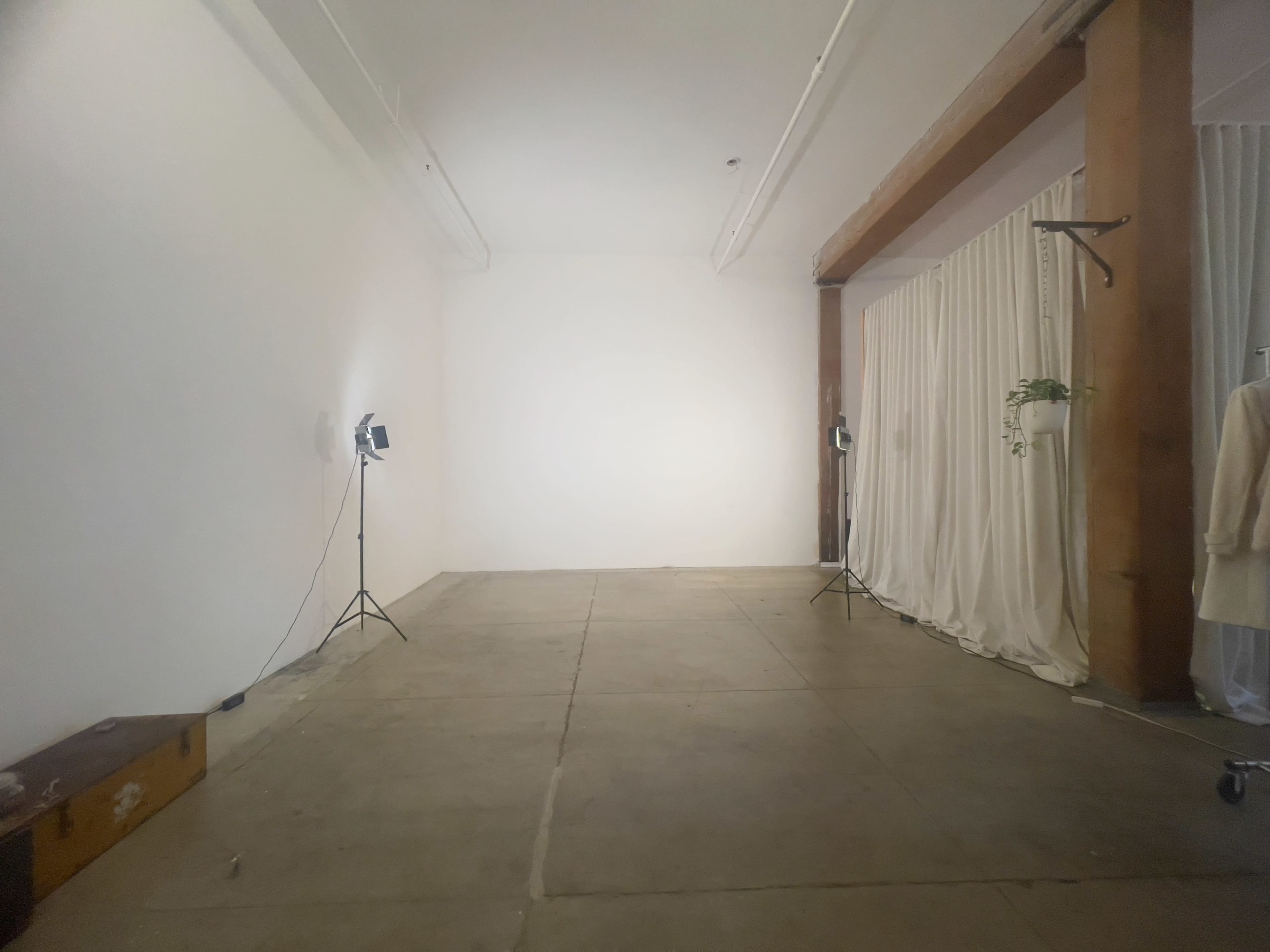An empty room features bare white walls, a concrete floor, and two light stands positioned on one side.