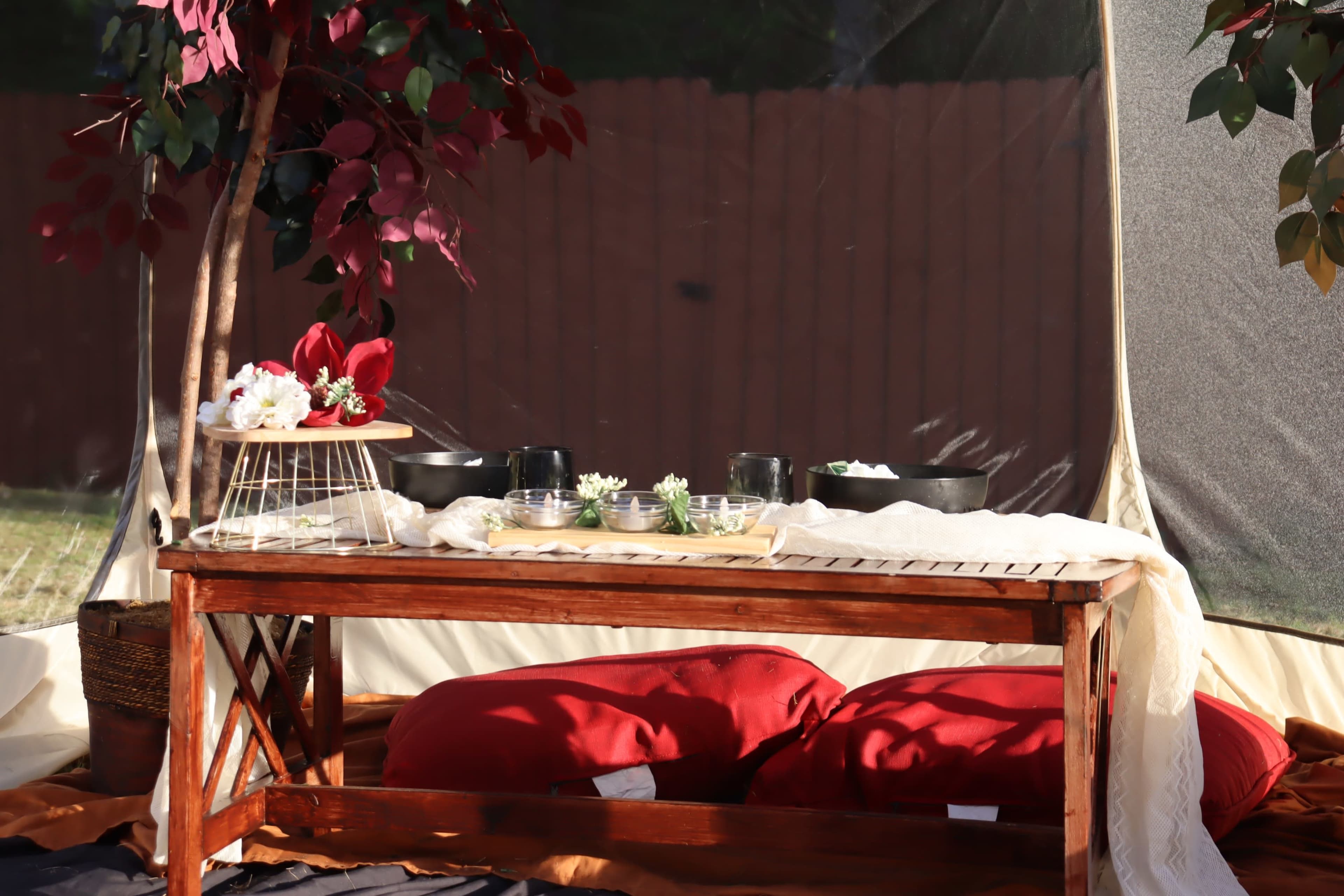 A wooden table is set up under a tent with various bowls and plants, surrounded by red cushions and a tree with maroon leaves.