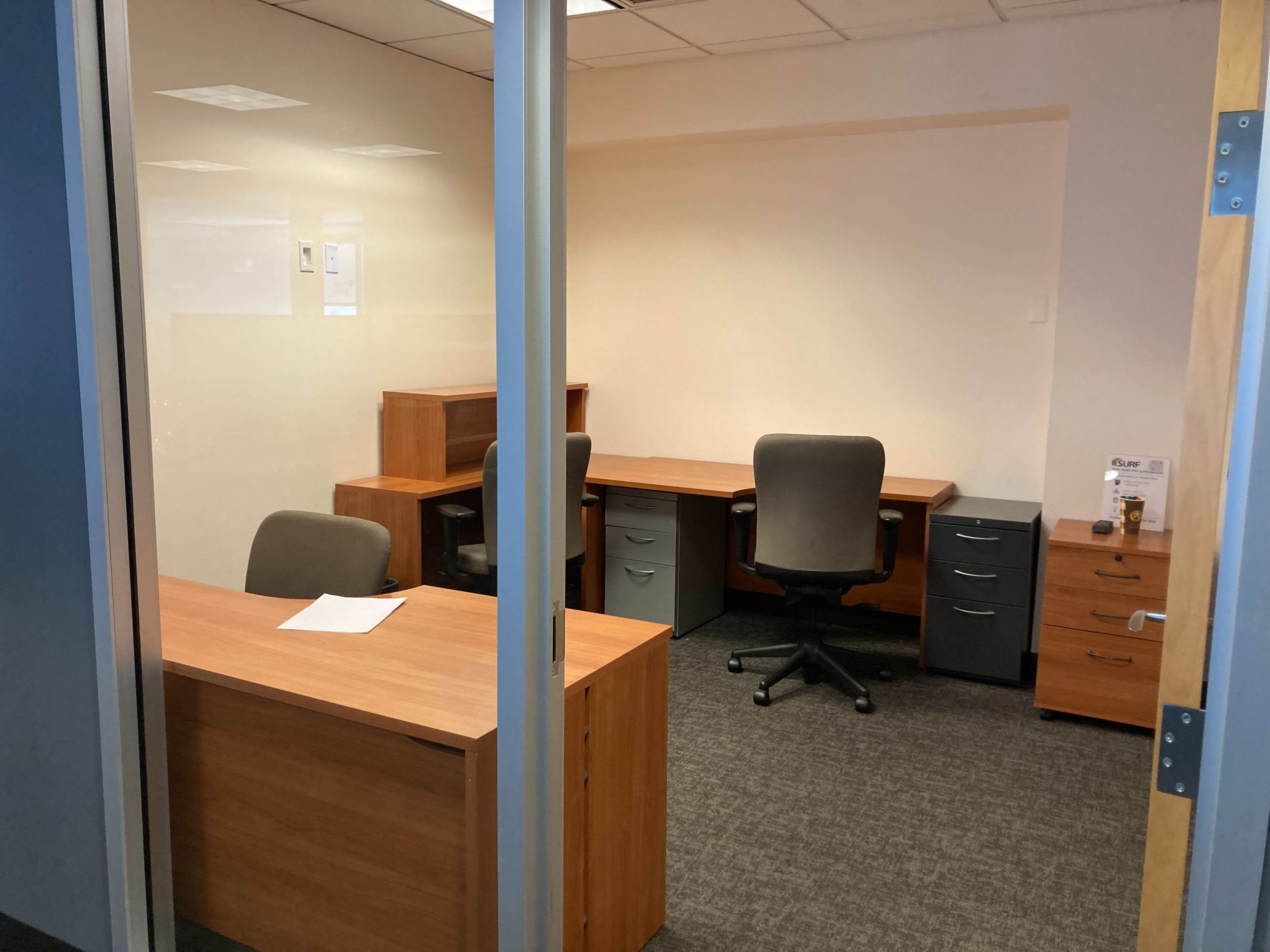 The image shows a simple office space with two wooden desks, grey filing cabinets, and chairs, all situated within a glass-walled room.