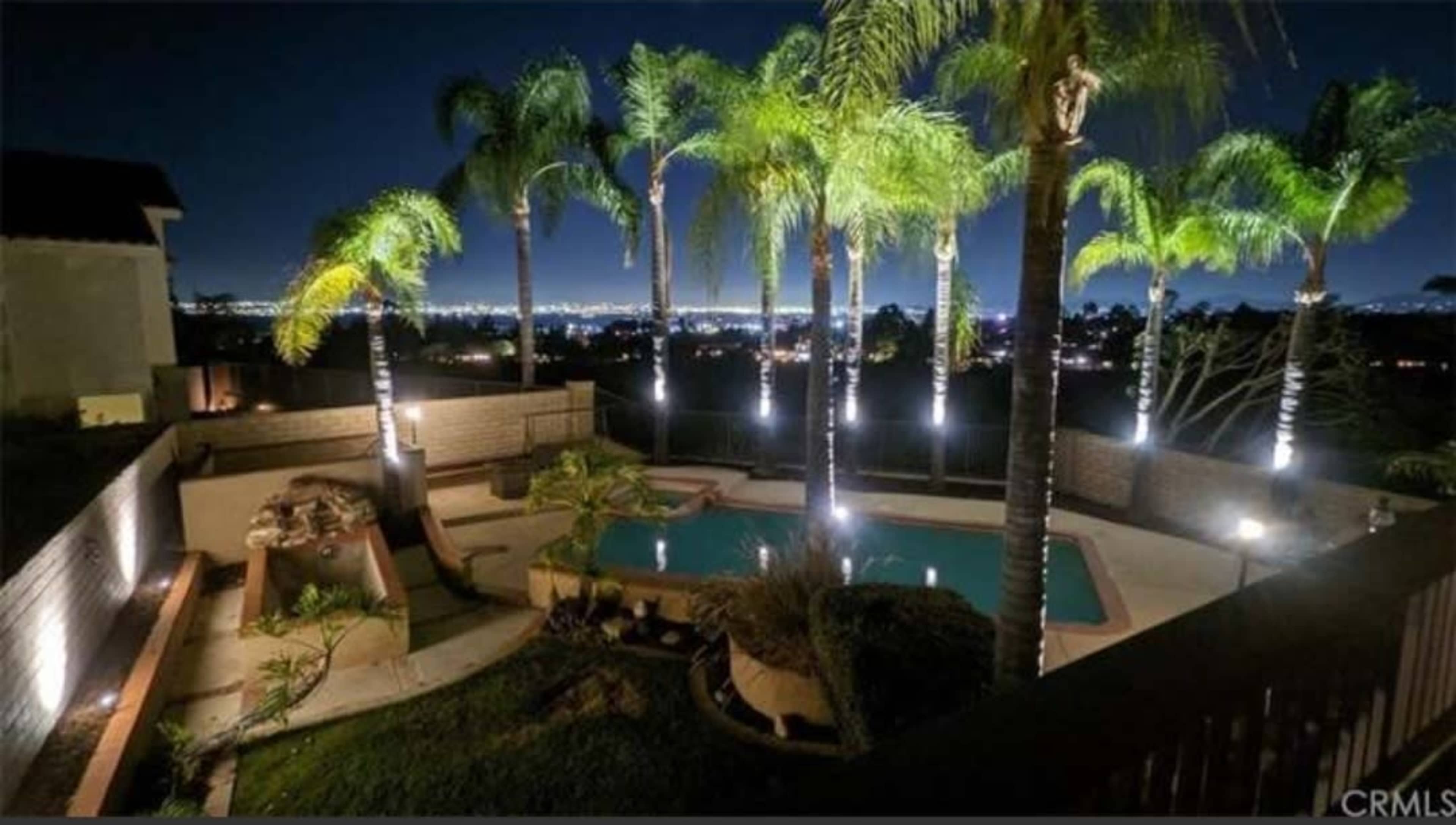 A backyard pool area surrounded by palm trees and illuminated by landscape lighting at night, with a view of a city skyline in the background.