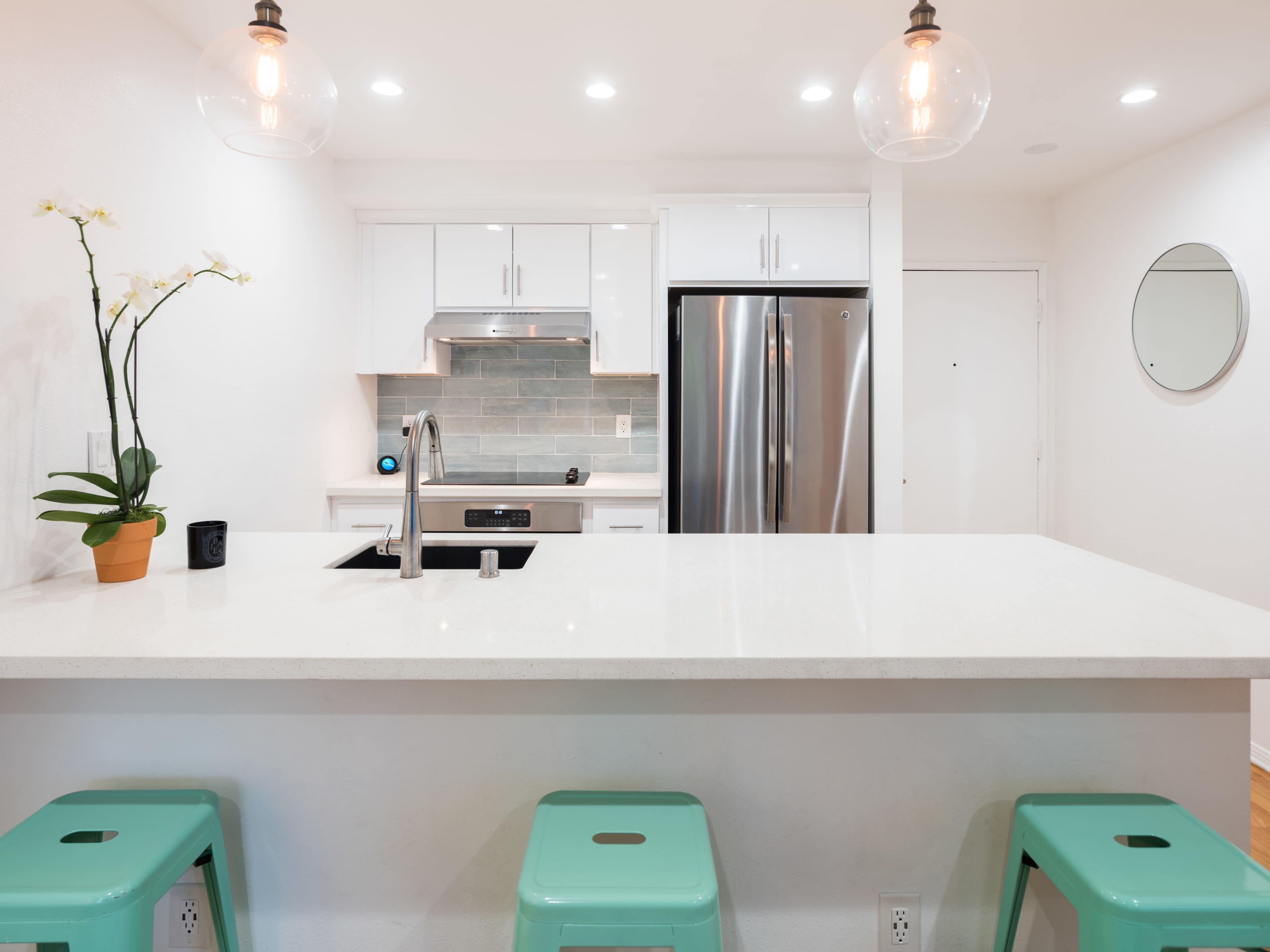 A modern kitchen features a white countertop island with turquoise stools, stainless steel appliances, and a sleek backsplash.
