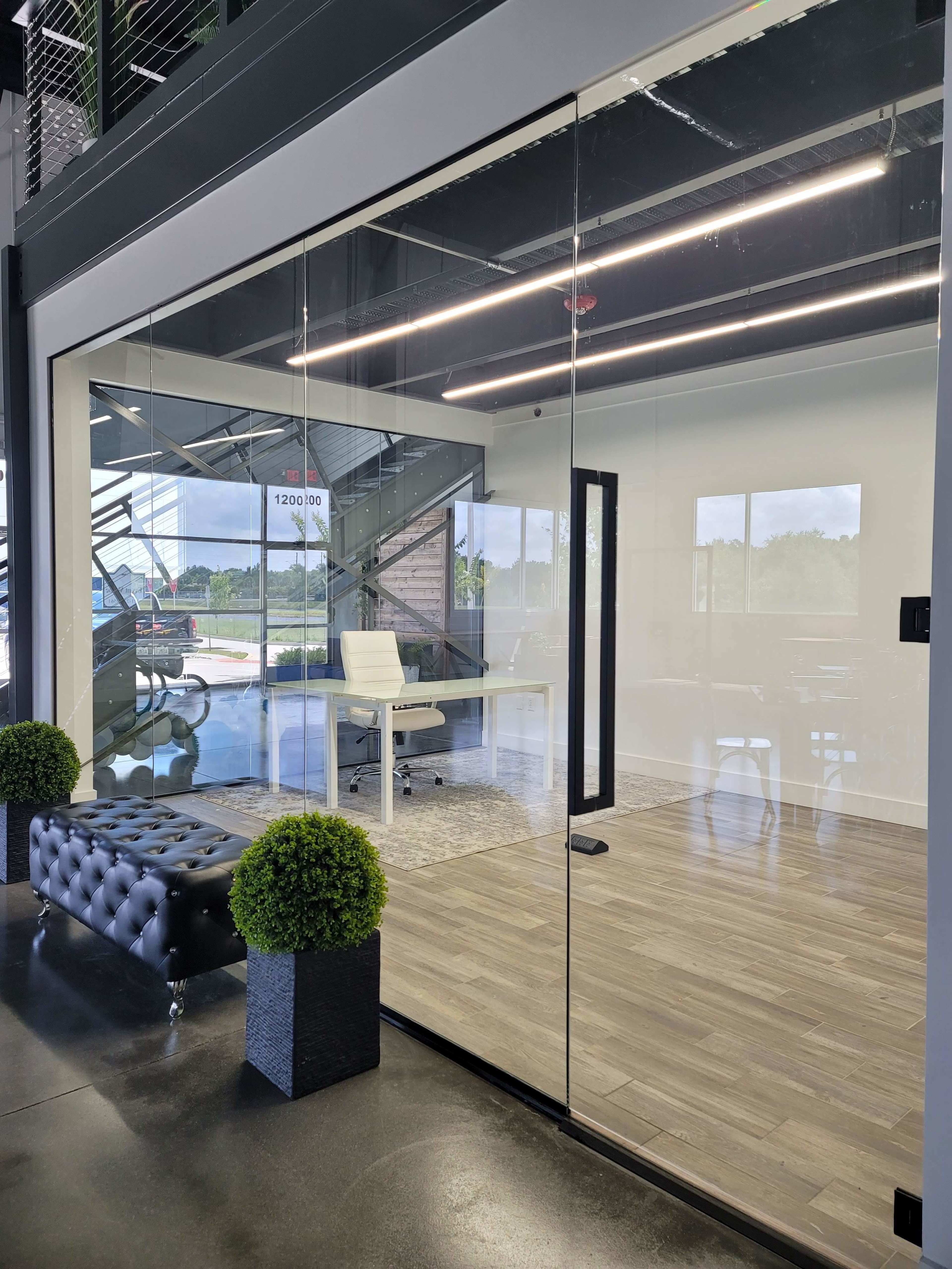 The image shows a modern office space with glass walls, featuring a sleek desk and chair inside, and two decorative plants flanking a black tufted sofa in the foreground.