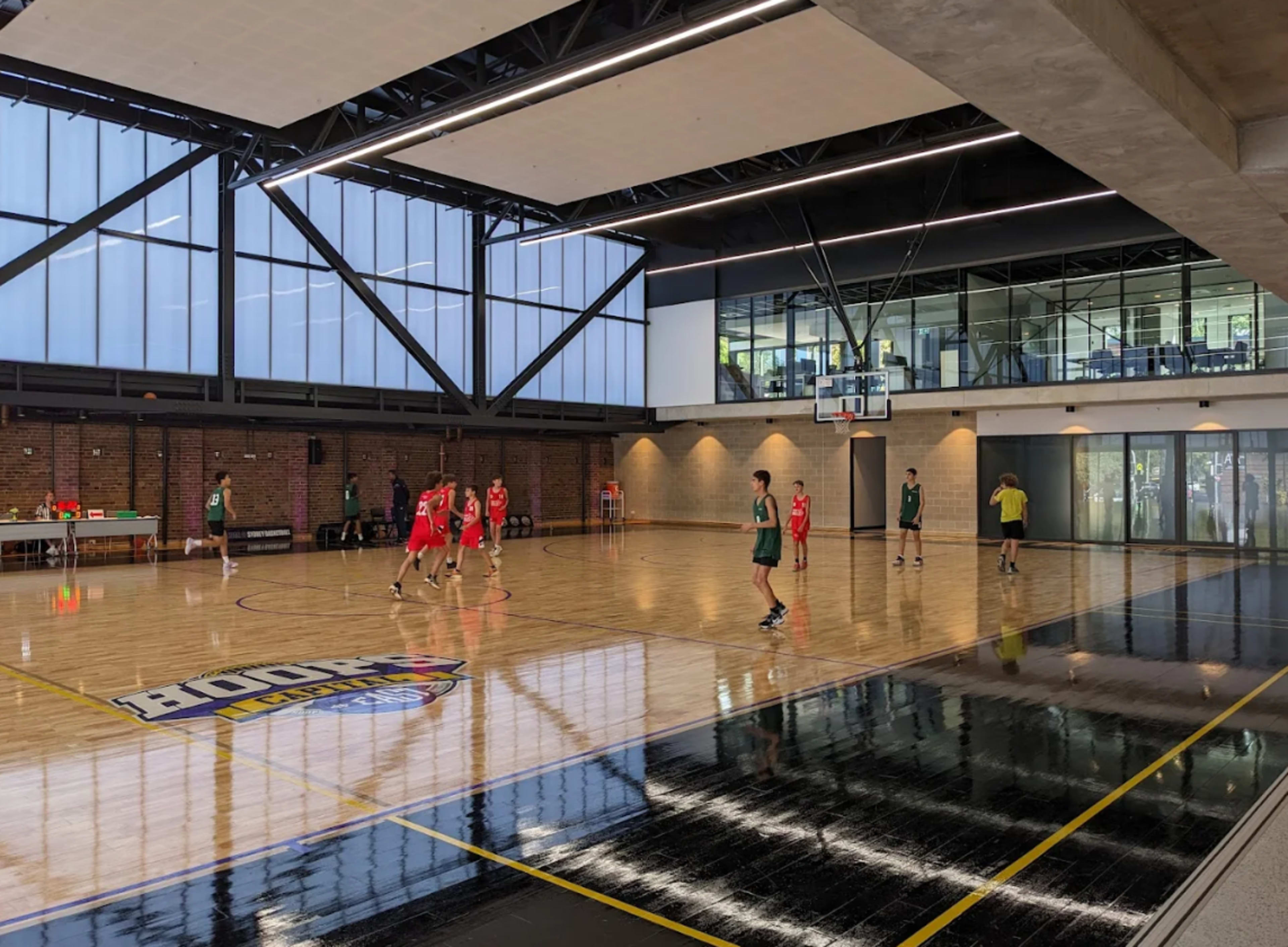 A basketball court features players in red and green jerseys engaging in a game, with large windows allowing natural light into the space.