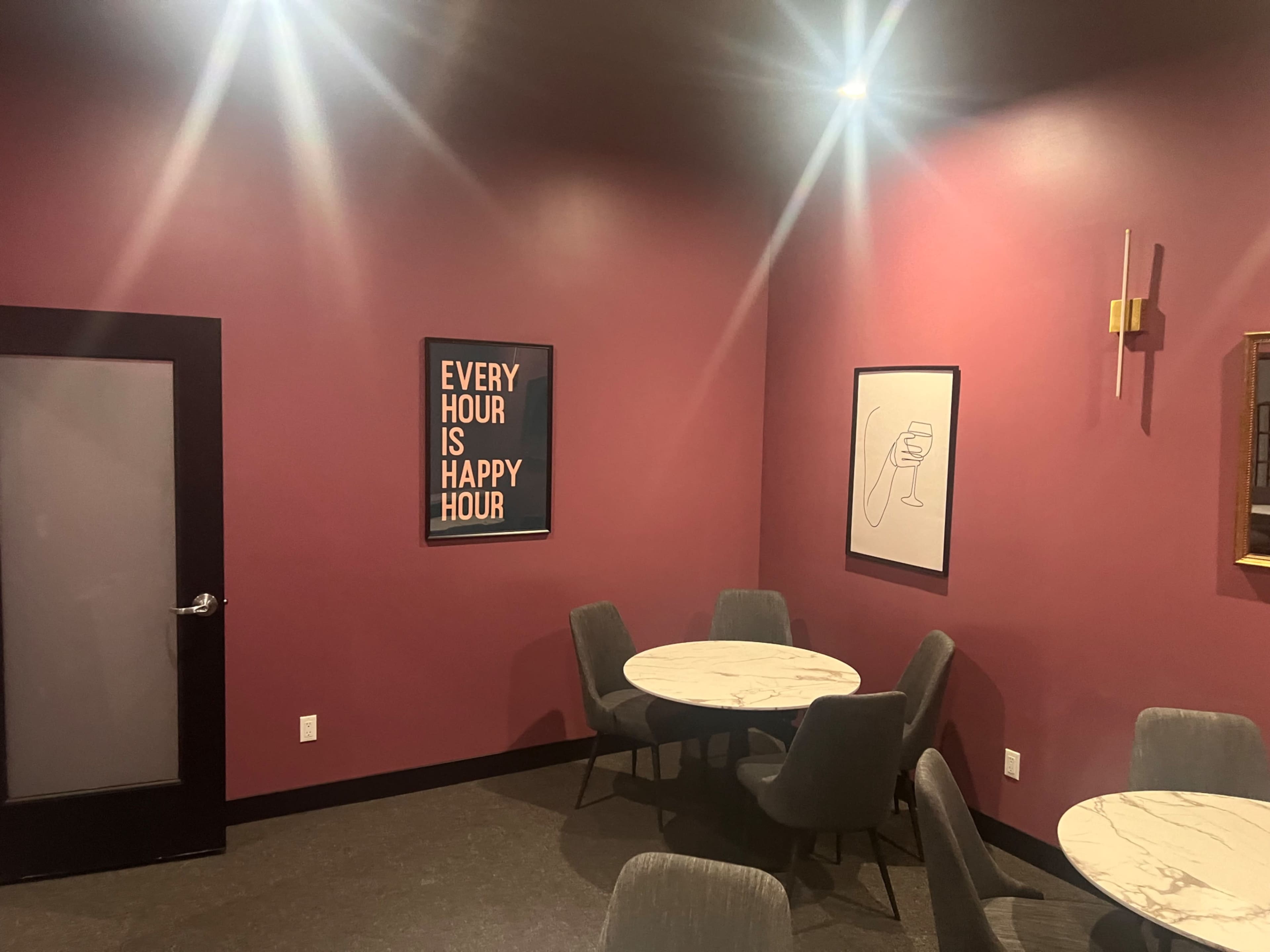 The image shows a dimly lit room with maroon walls, featuring marble-topped tables and a few chairs, along with framed artwork on the walls.