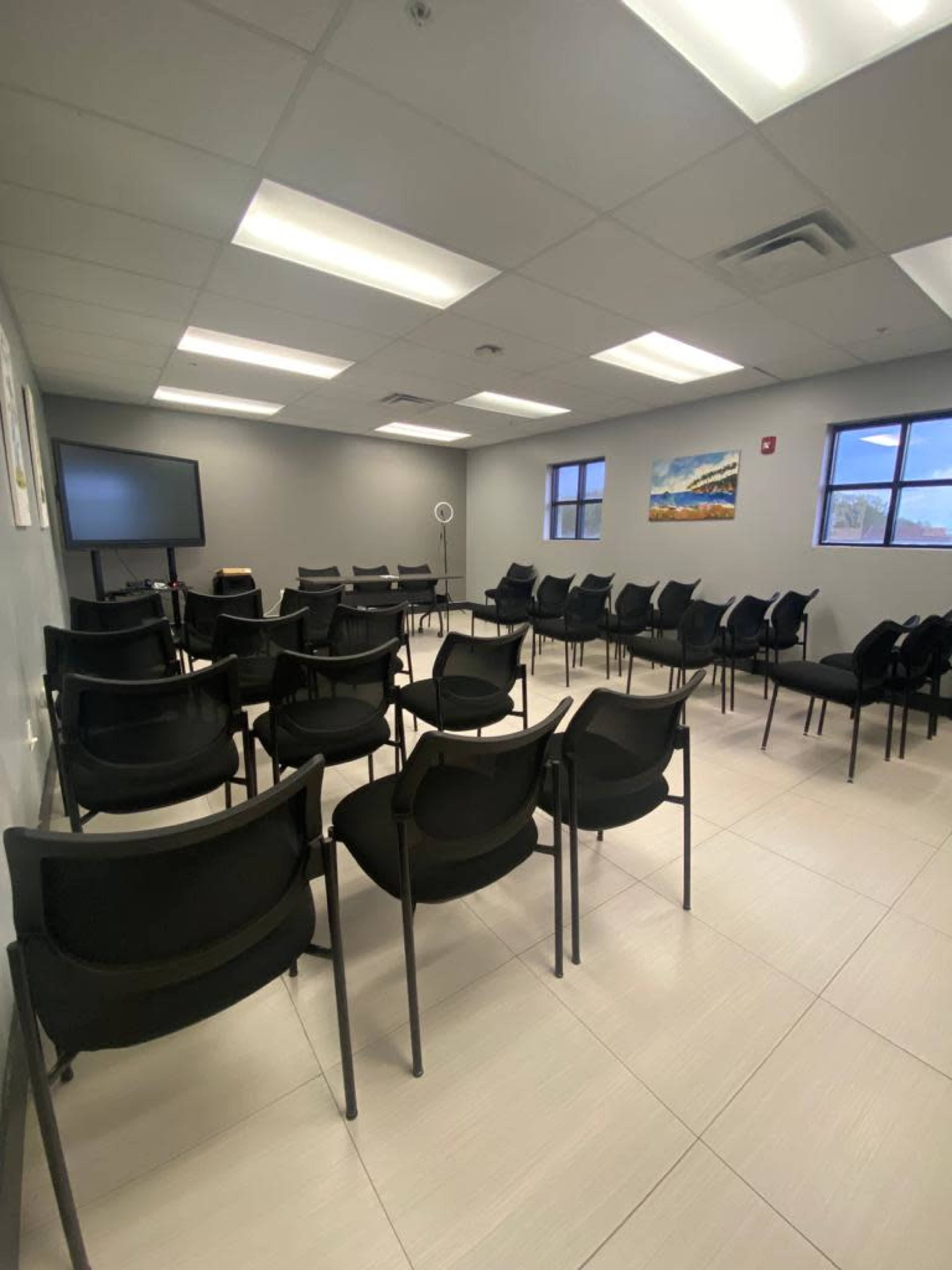 A waiting room with multiple black chairs arranged in rows, a television screen at the front, and windows on one side.